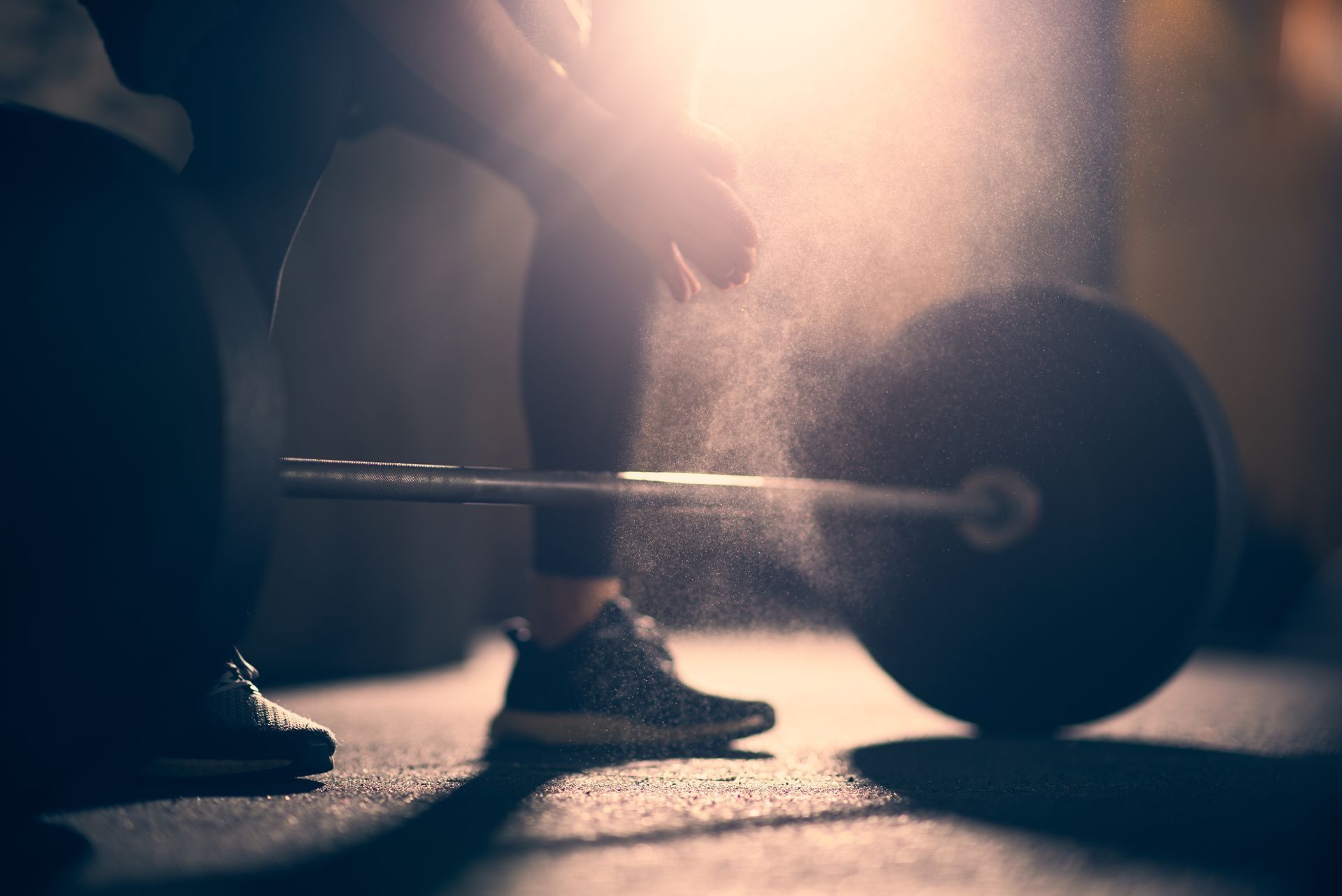 Person lifting a barbell in a dim gym, backlit by warm light.