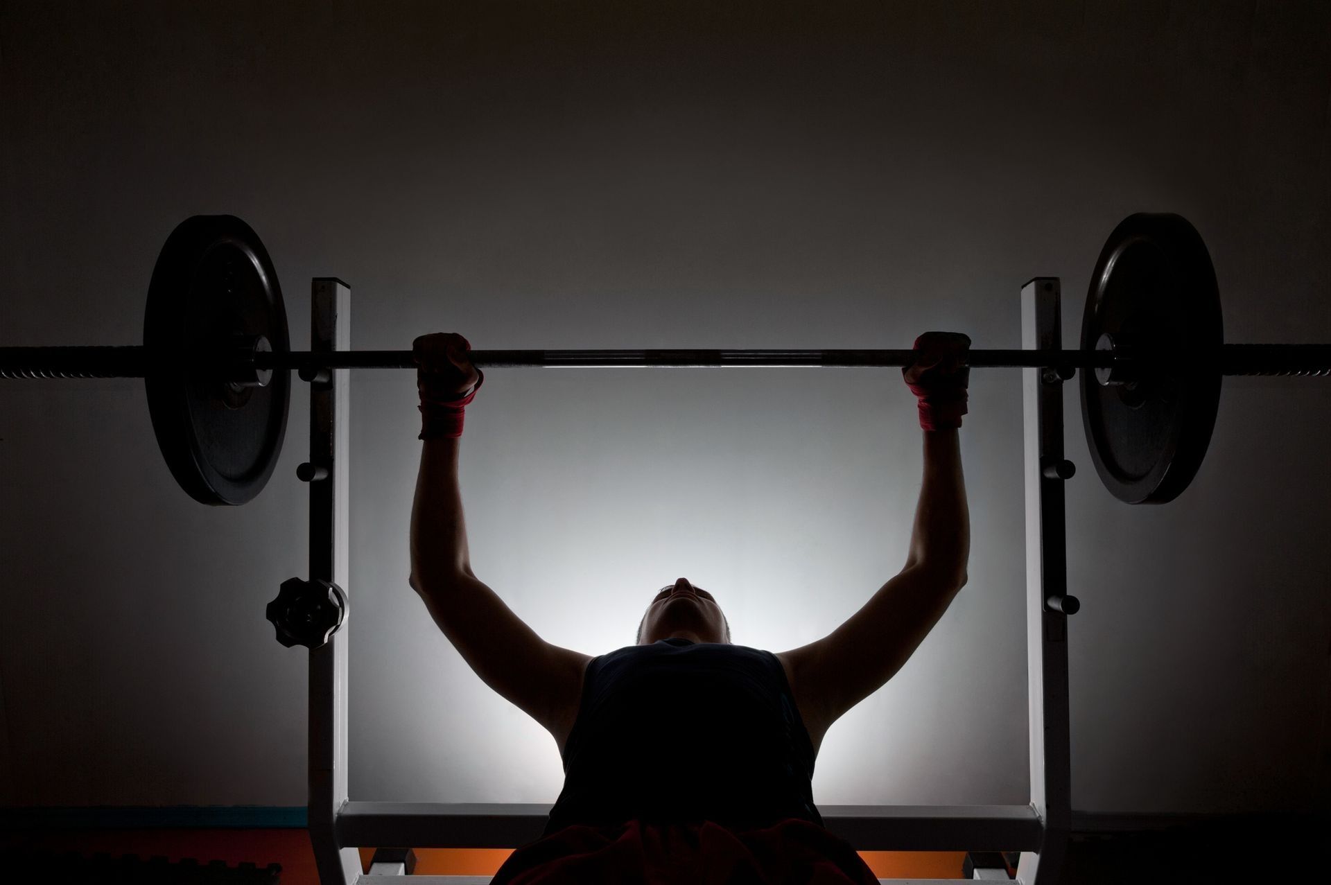 Person bench pressing a barbell in a dim gym, silhouetted against bright backlighting