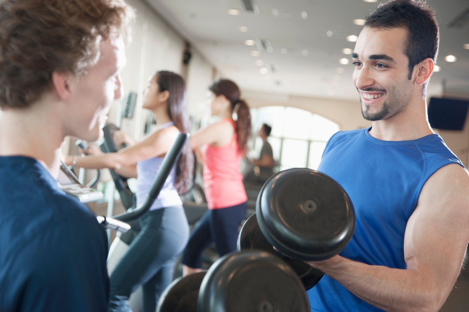 People lifting dumbbells in a gym, with a smiling man in a blue shirt in the foreground