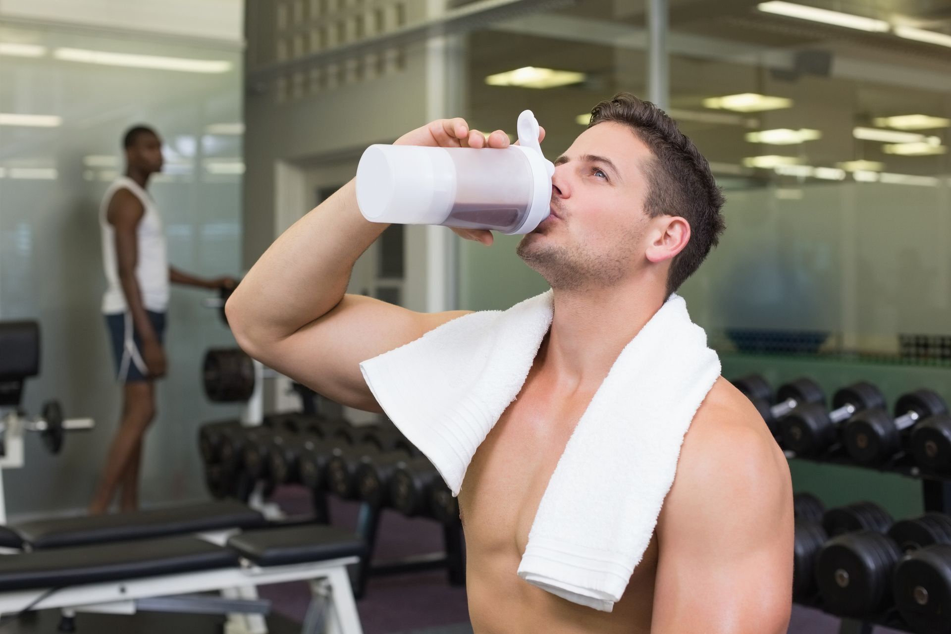 Man drinking water with a towel around his neck in a gym, dumbbells and another exerciser in the background
