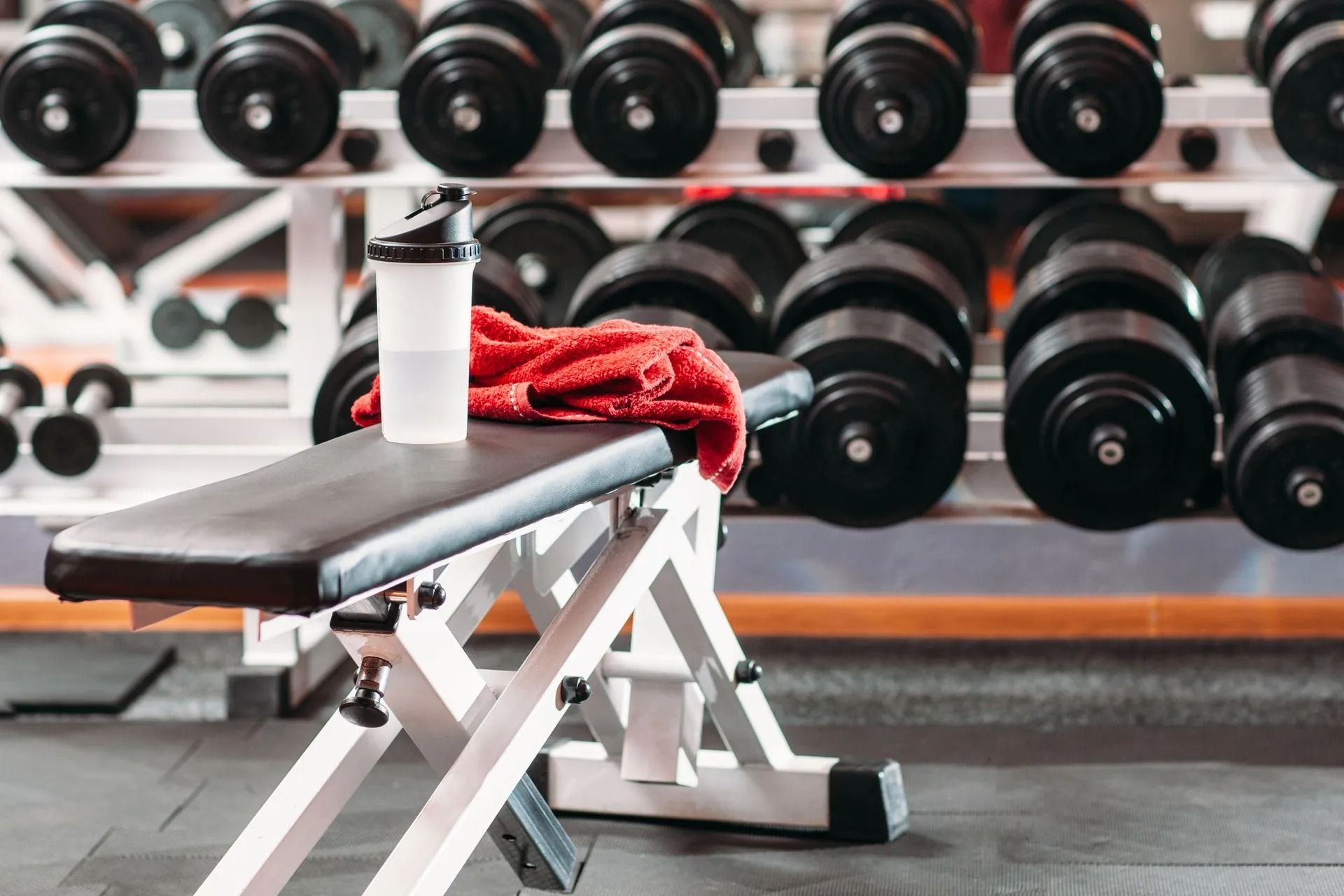Gym bench with a water bottle and red towel in front of dumbbell racks