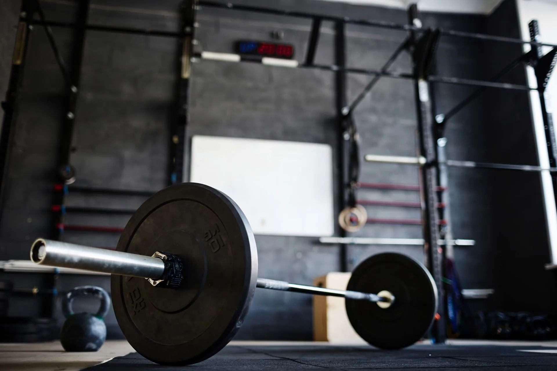 Barbell with weight plates on a gym floor, with a kettlebell and wall-mounted pull-up rig in the background