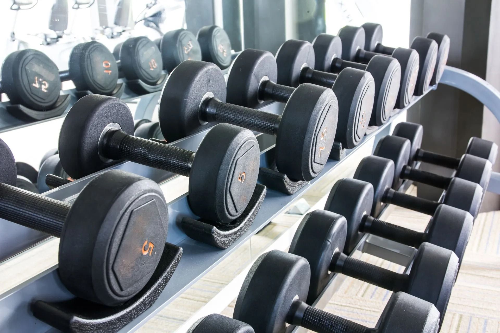 Rows of black dumbbells on a gym rack beside a mirror