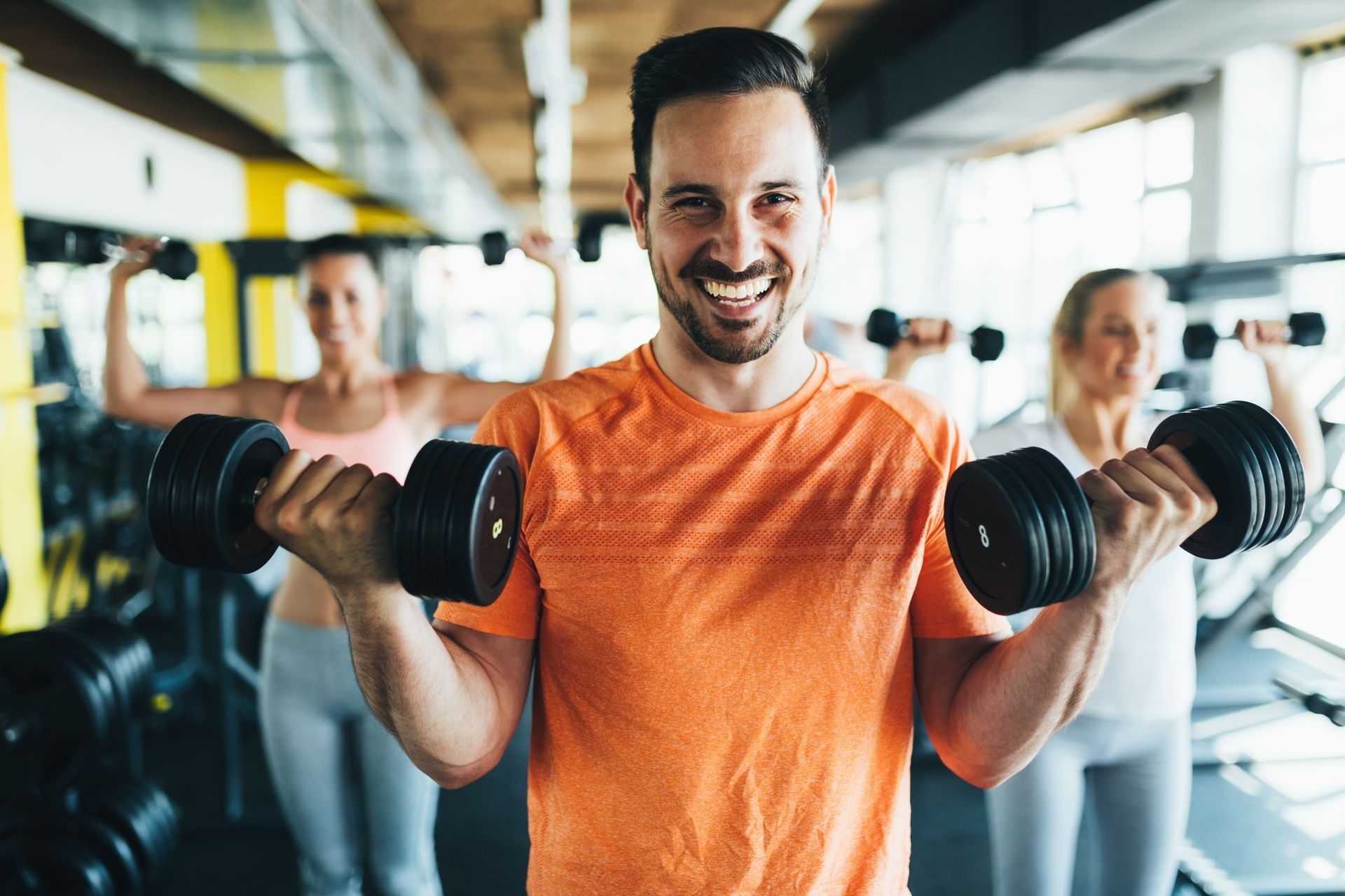 Man in orange shirt lifting dumbbells in a gym, smiling, with other exercisers in the background