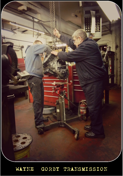 Two men working on a transmission in a repair shop. One man uses a chain hoist. Tools and red tool chest visible.