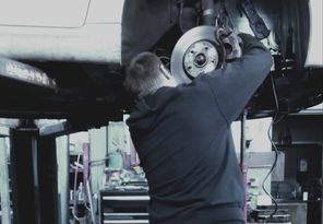 Mechanic working on a car's brake disc while the car is raised on a lift in a garage.
