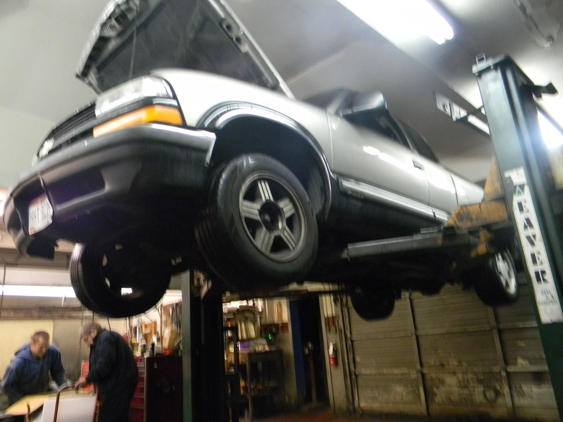 Silver truck on a lift with hood open inside auto repair shop. Two mechanics working nearby.