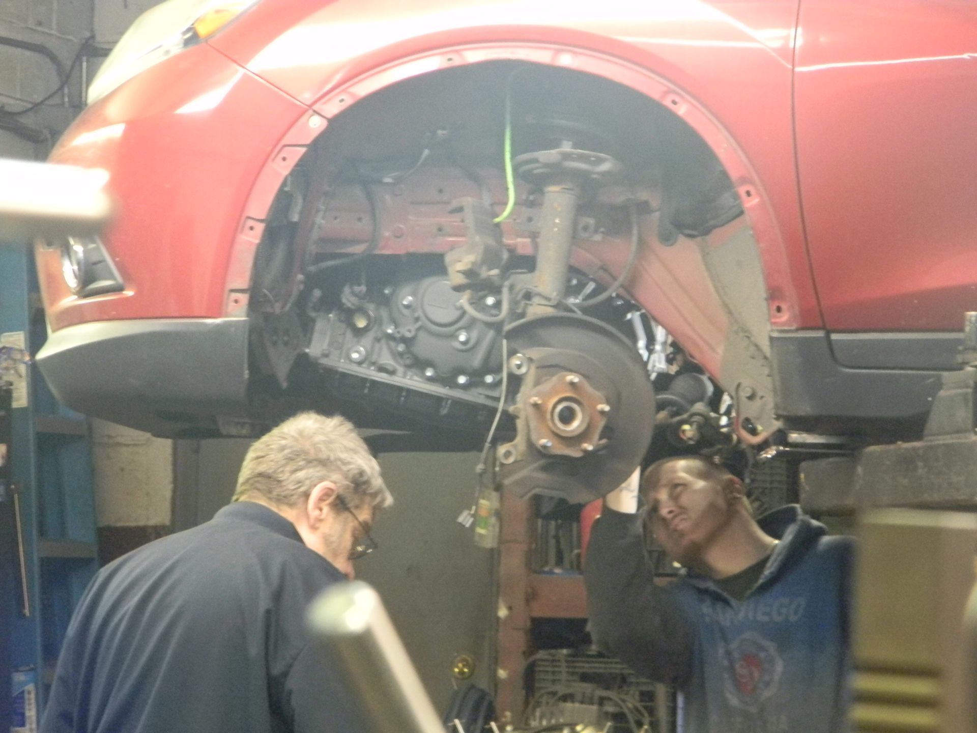 Two people working on the front wheel assembly of a red car in a garage. One person is looking down, the other is holding the assembly.