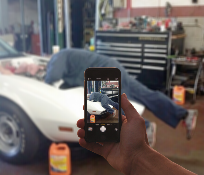 A person uses their phone to photograph a mechanic working on a white car in a garage.