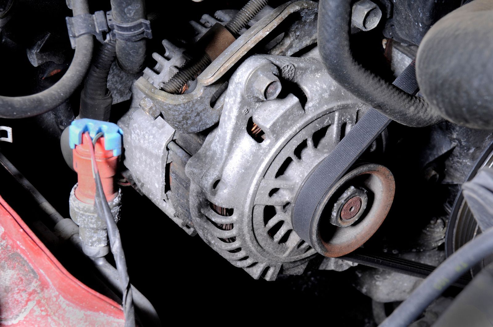 Close-up of a car alternator with a belt and various connected wires and tubes in an engine bay.