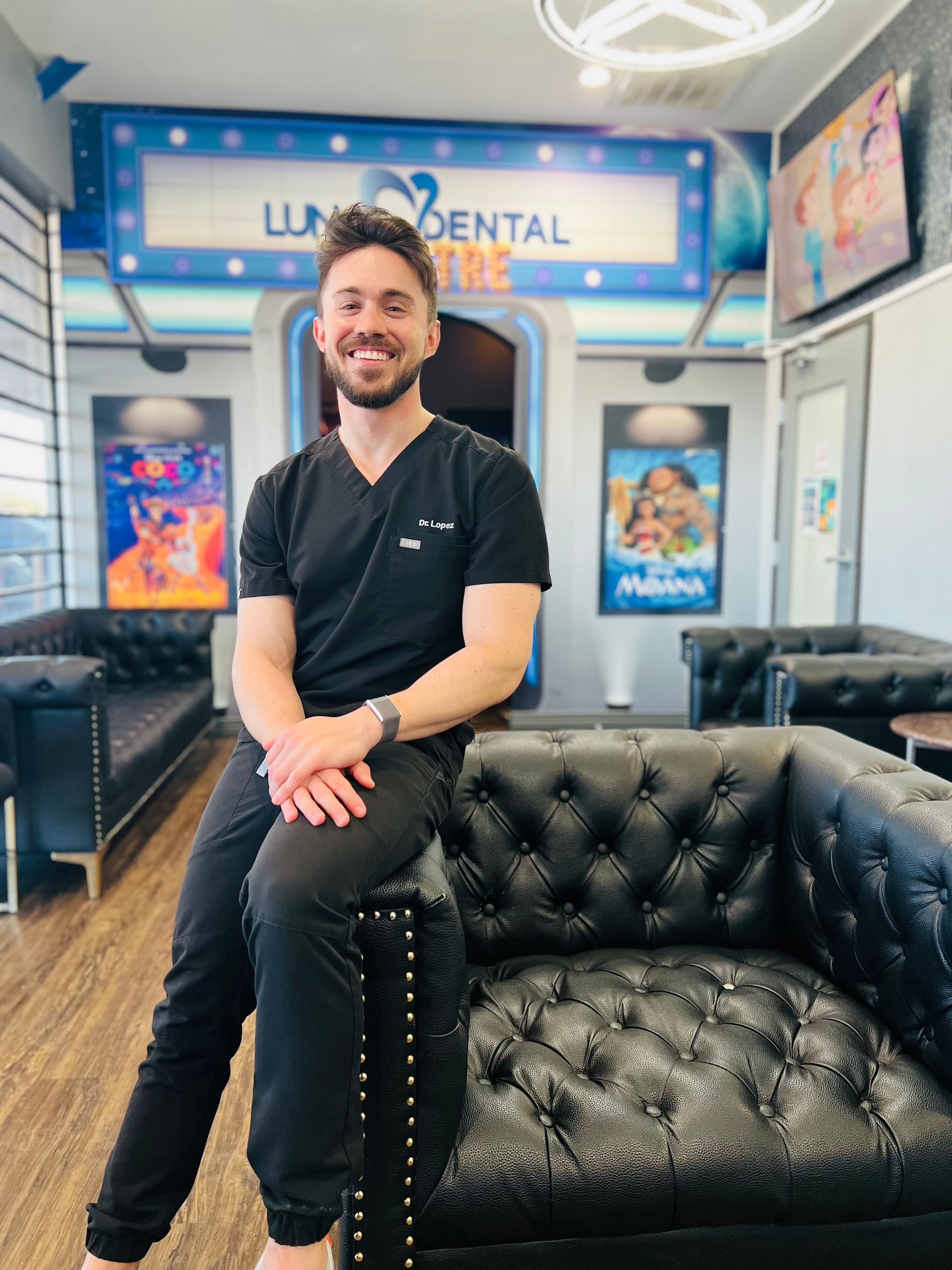 Luna-Dental-Dallas- Man in black scrubs sits on a black leather couch in a dental office, smiling.