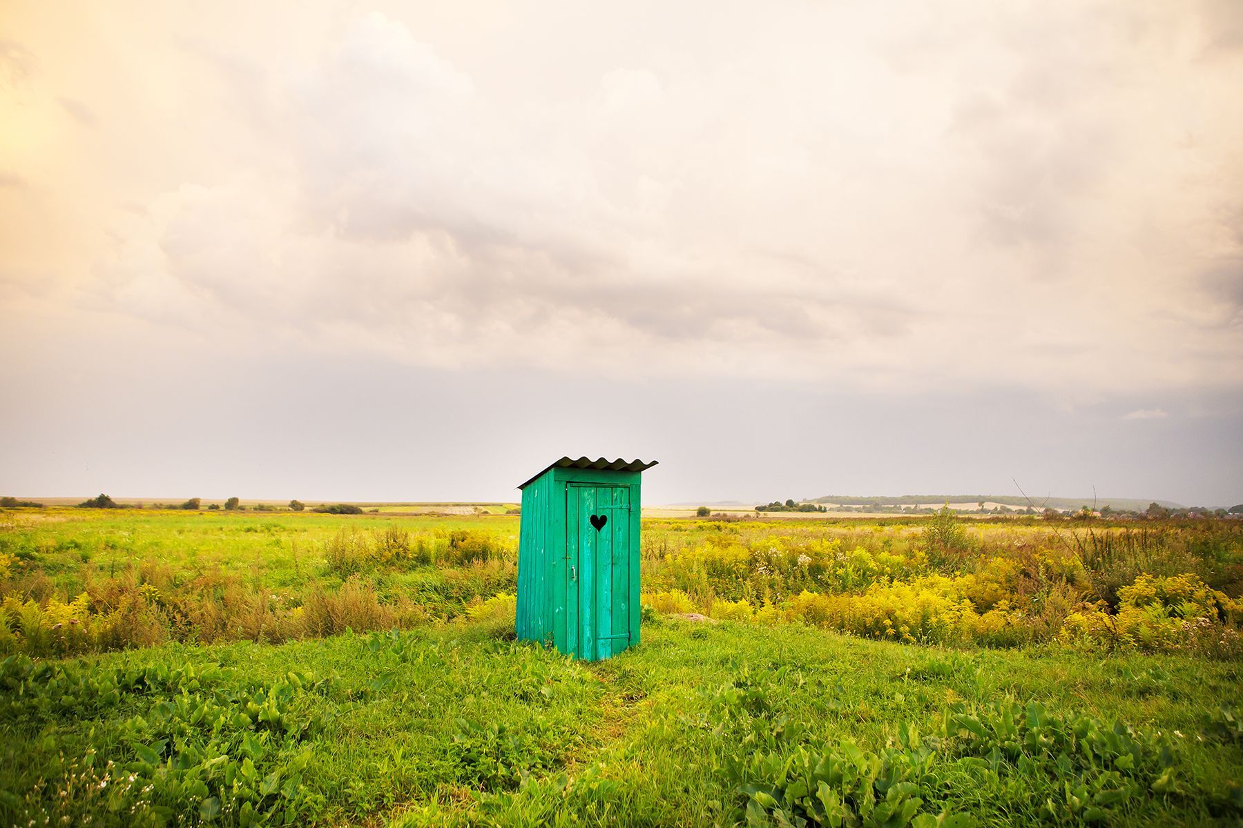 A green outhouse is sitting in the middle of a grassy field.