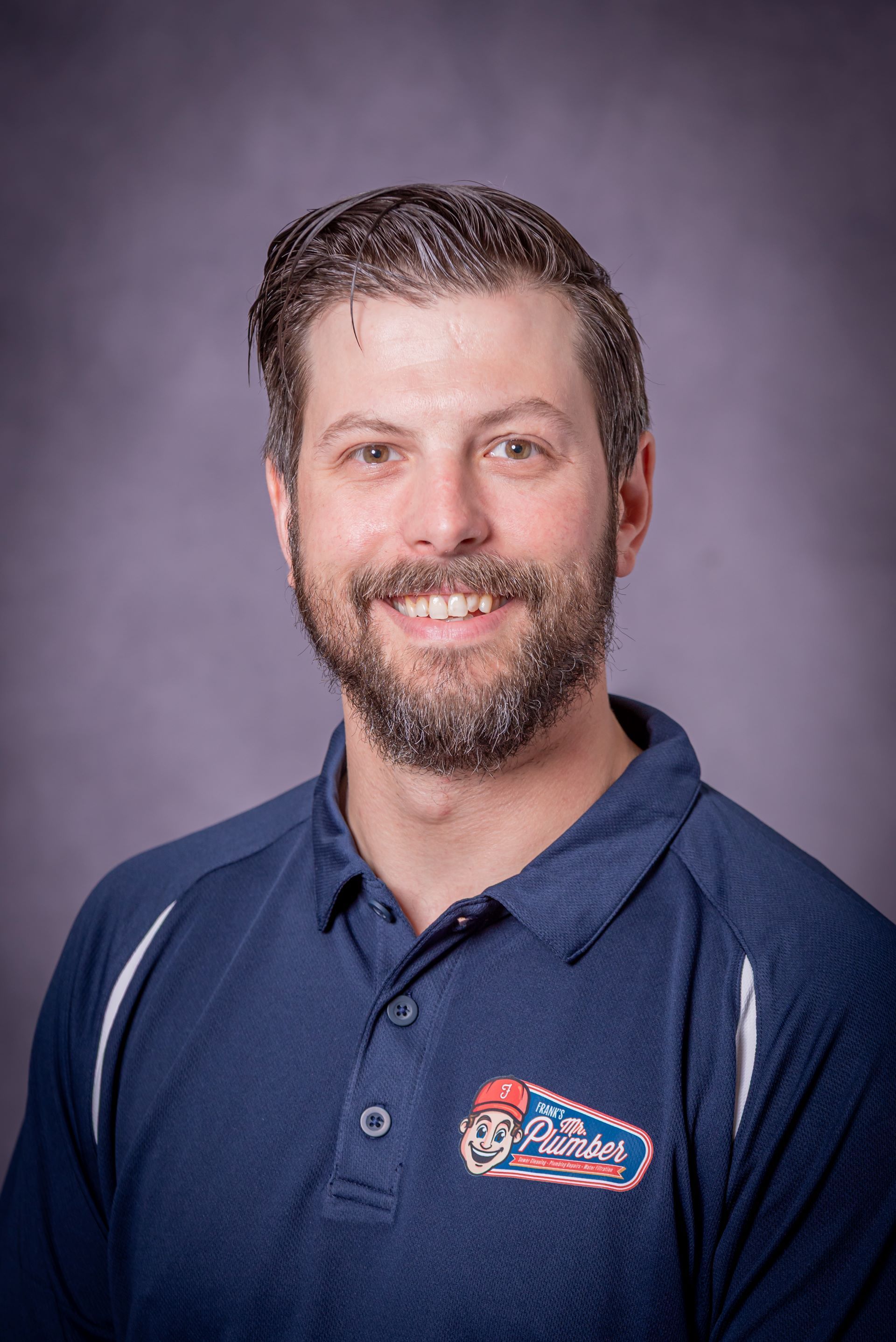 A man with a beard is wearing a blue polo shirt and smiling.