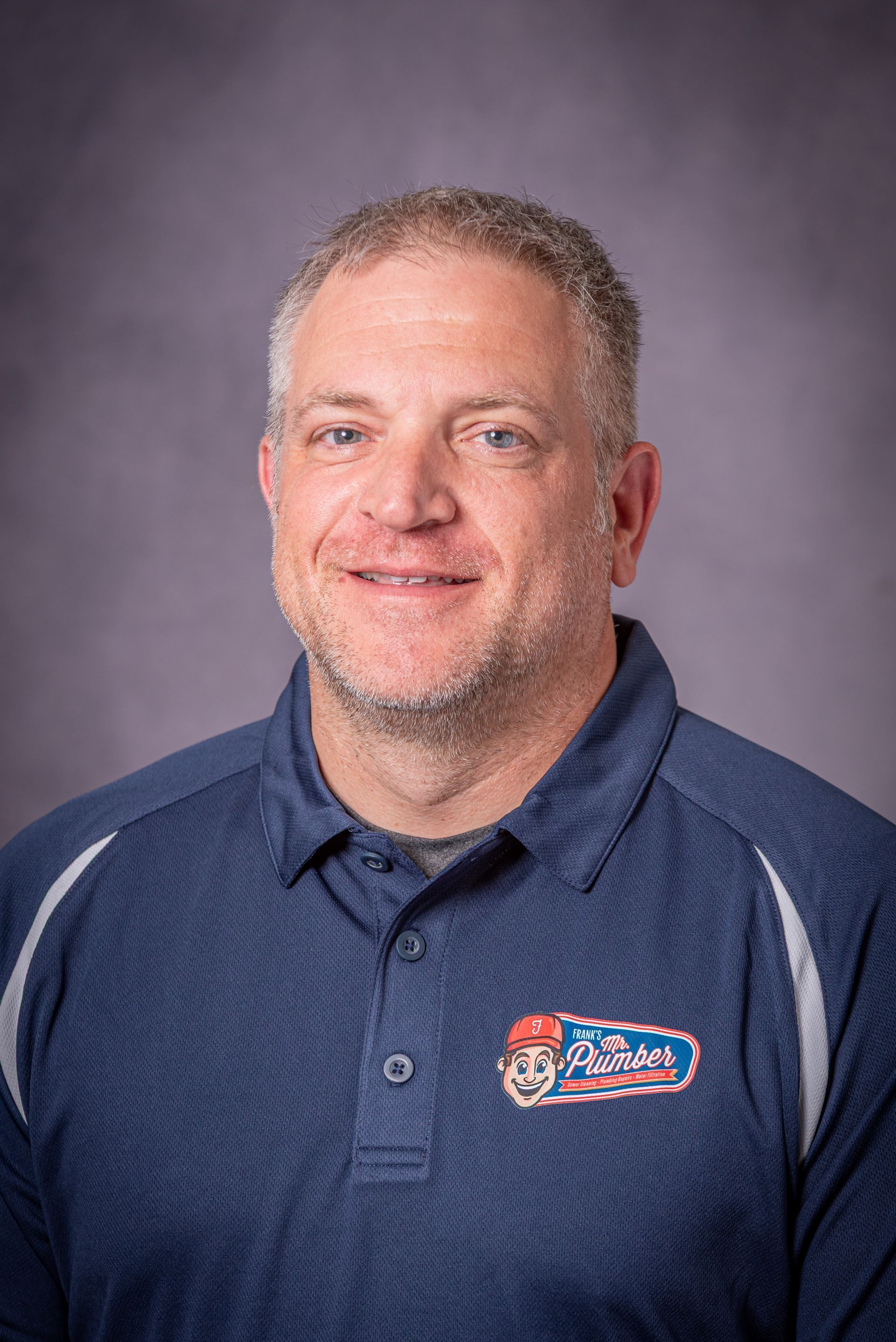 Man in a blue polo shirt, smiling, with a logo on the left chest, posing against a gray background.