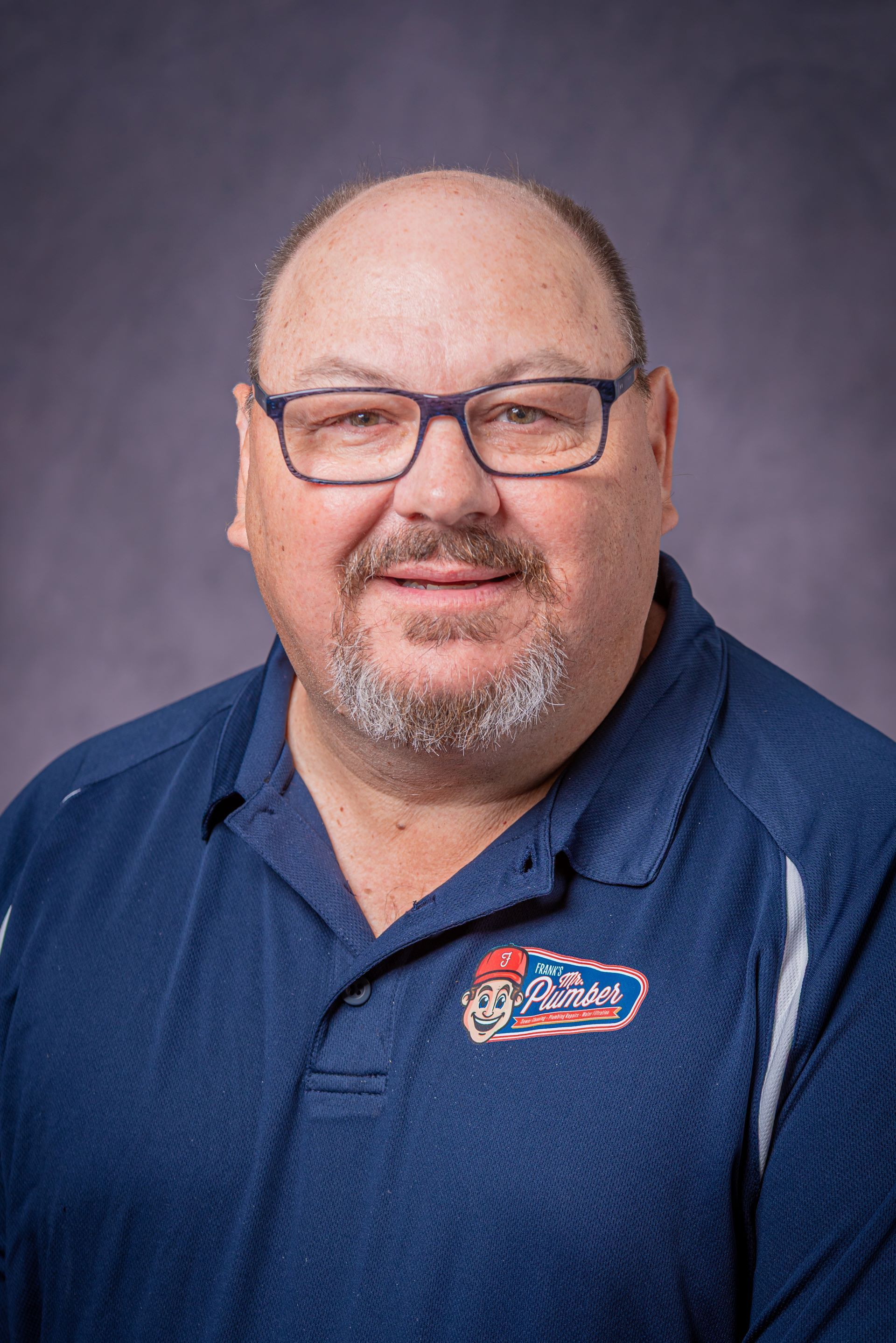 Man with glasses and goatee wearing a blue shirt with a logo, posing in front of a gray backdrop.