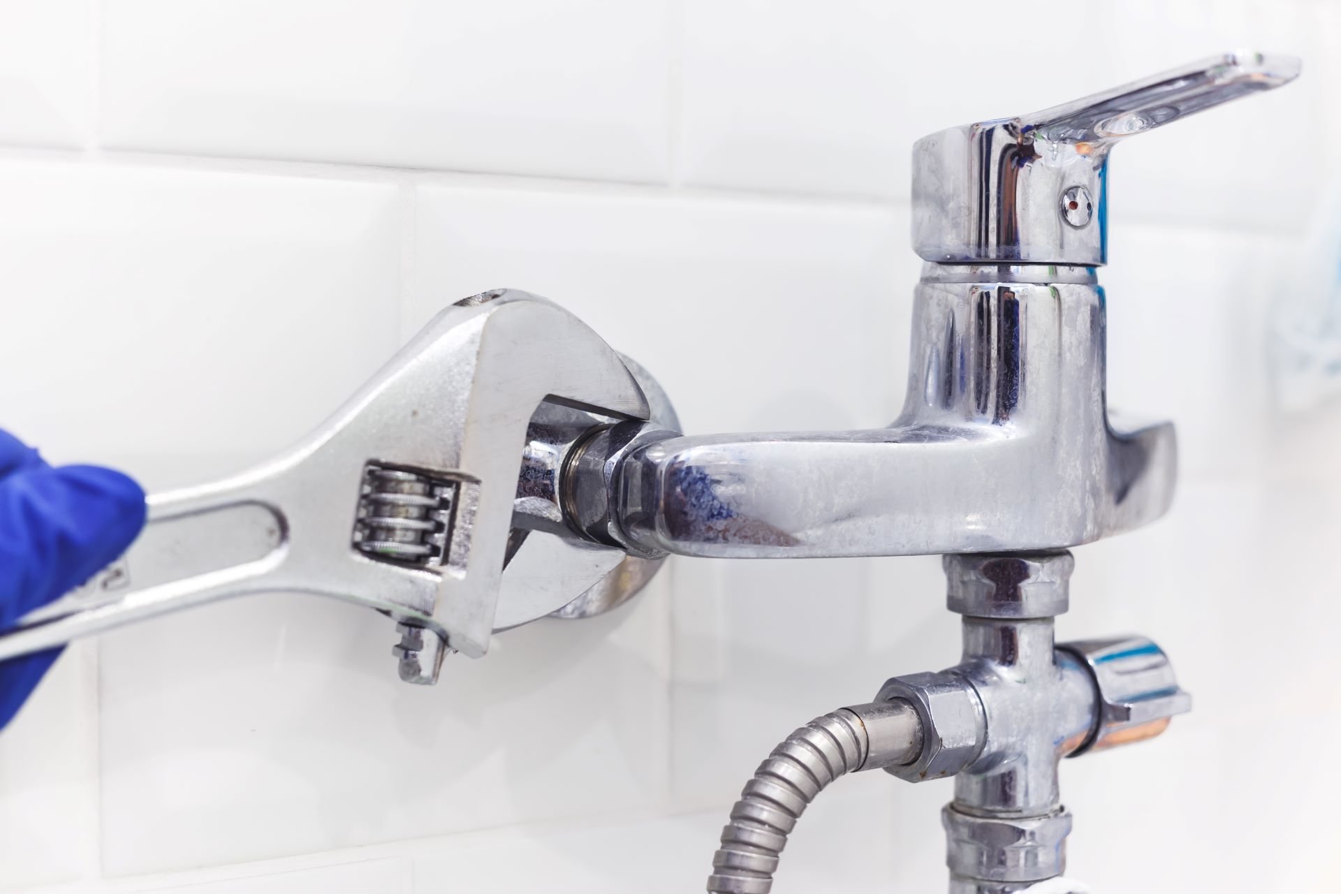 A person with a wrench tightens a faucet on a white tiled wall.