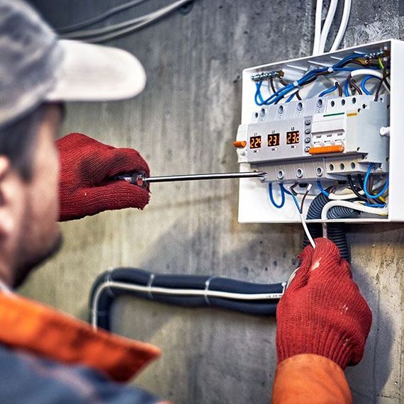 Electrician wiring a wall-mounted electrical panel with a screwdriver and red gloves