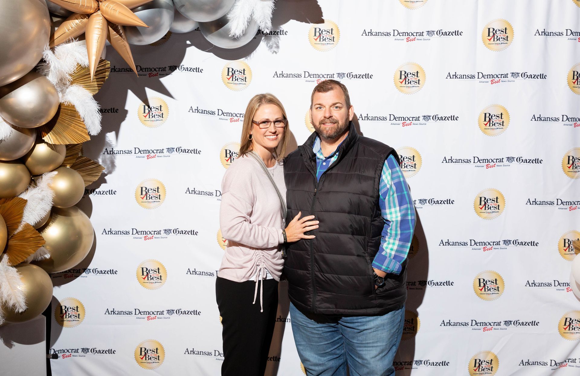 A man and a woman are posing for a picture in front of a backdrop with balloons.