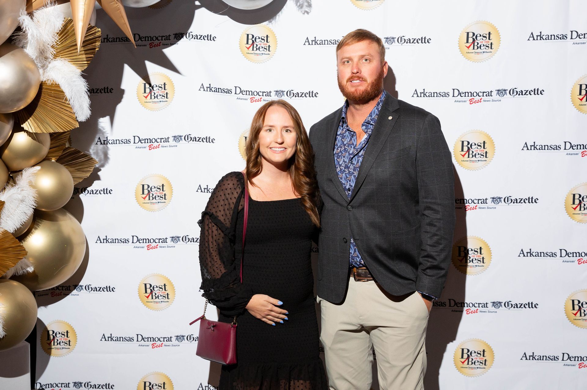 A man and a woman are posing for a picture in front of a wall with balloons.