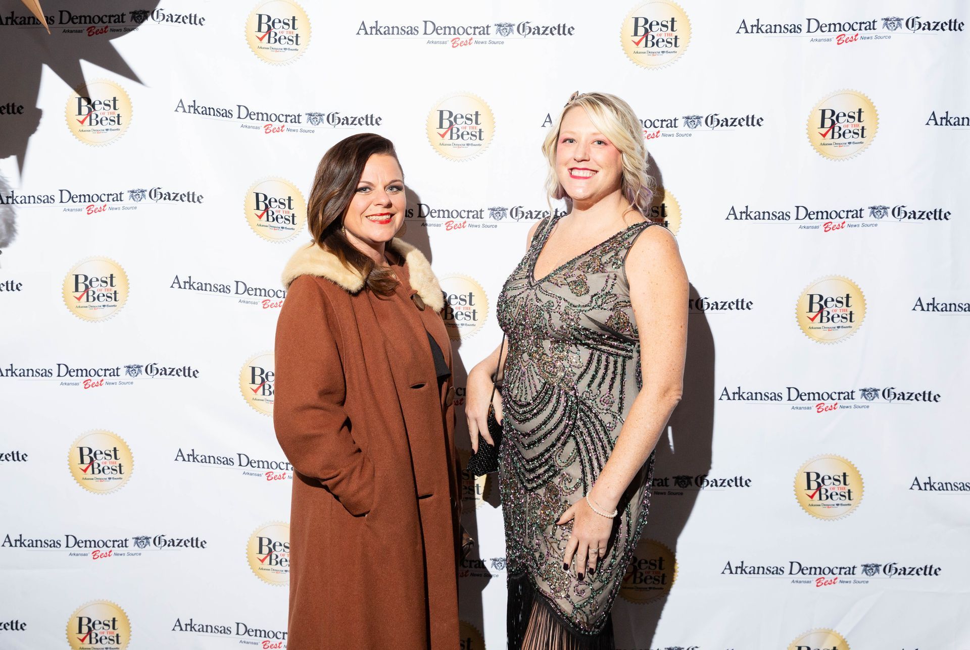 Two women are standing next to each other in front of a white wall.