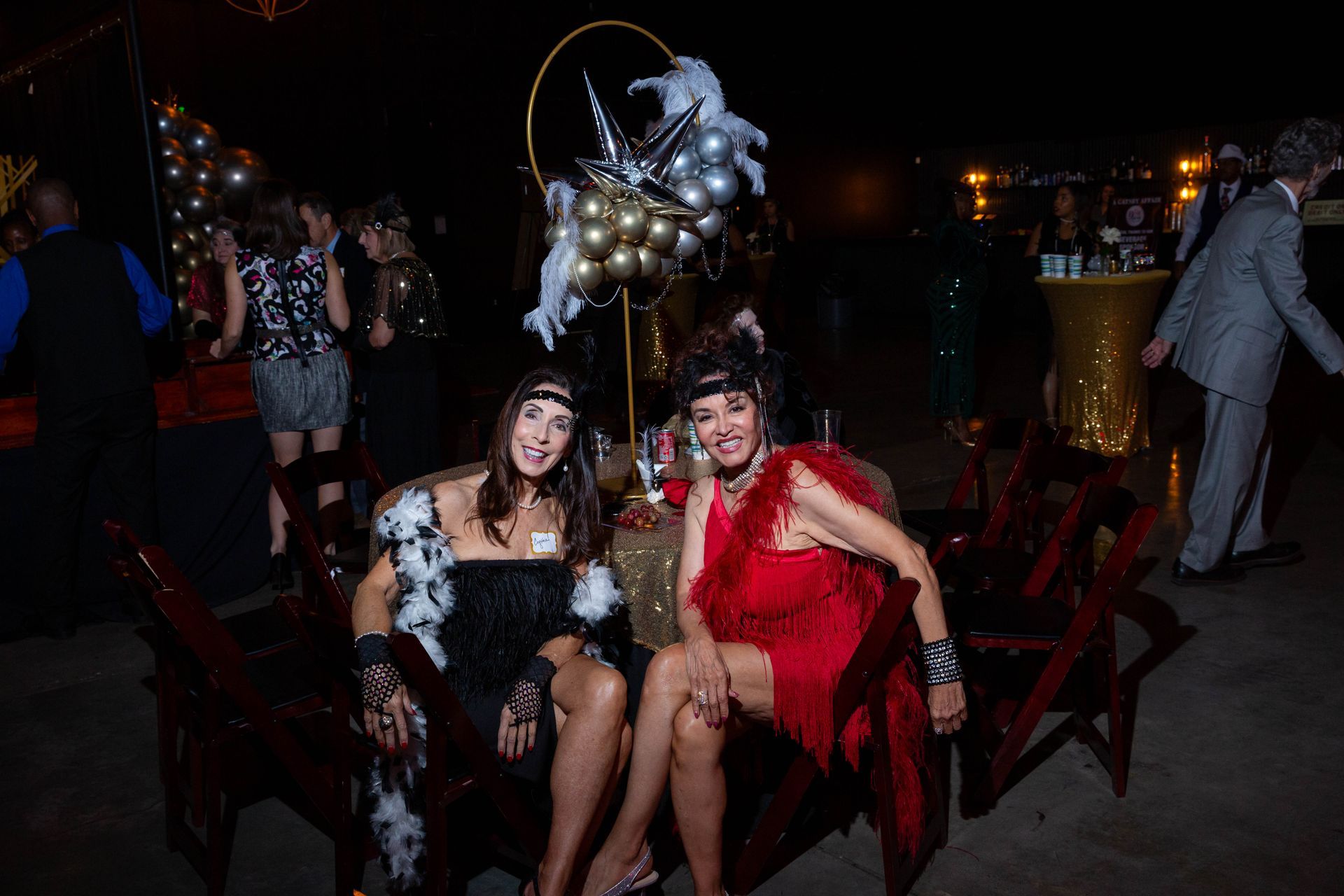 Two women are sitting next to each other in a chair at a party.
