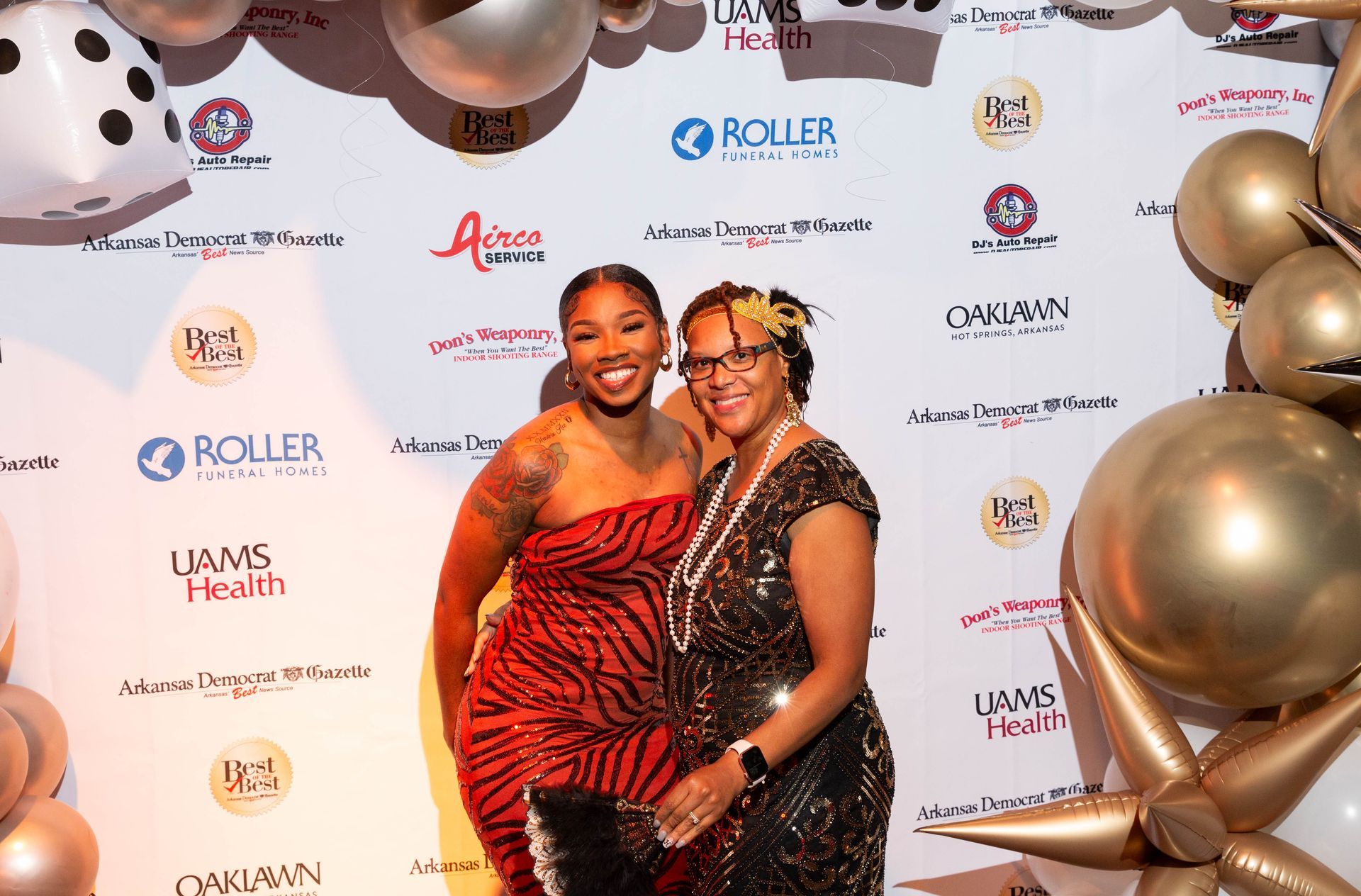 Two women are posing for a picture in front of a wall with balloons.
