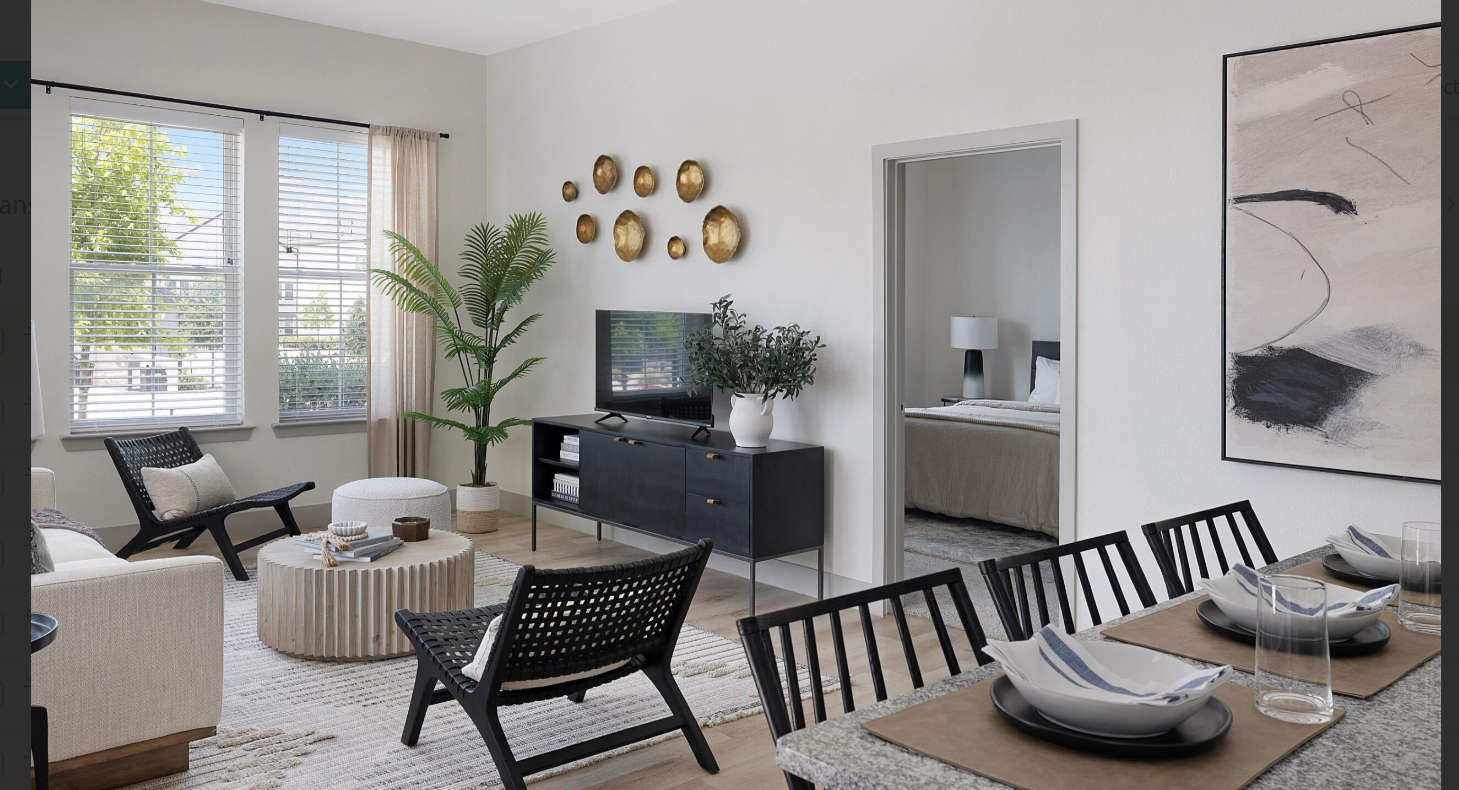 Bright living-dining room with black chairs, a round coffee table, wall art, and a bedroom visible through an open door.