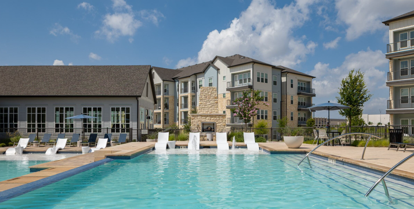 Resort-style outdoor pool with lounge chairs and a modern apartment building under a blue sky.