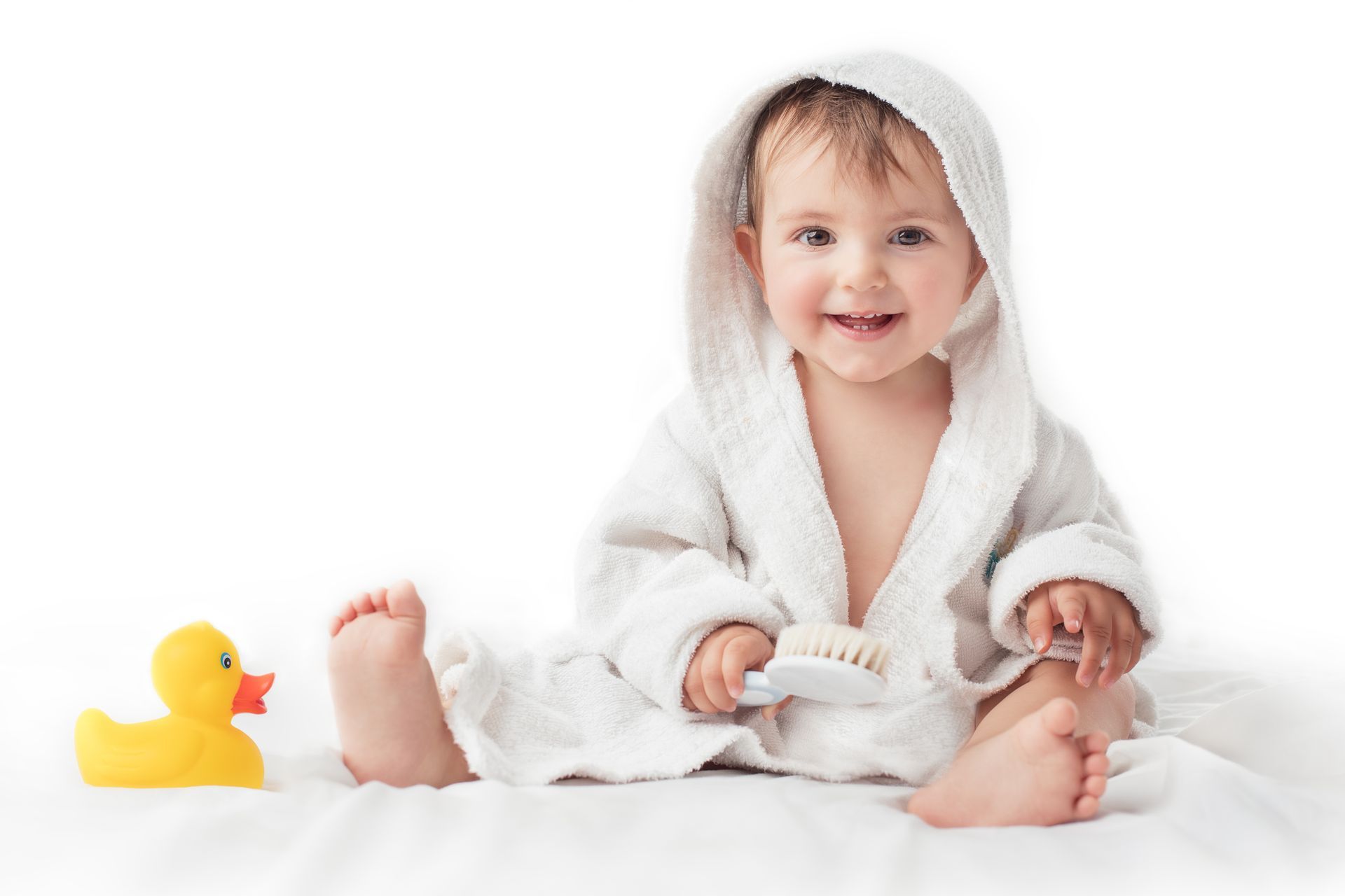 A baby is sitting on a bed with a rubber duck and a brush.
