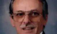 Man with mustache, glasses, and dark blazer smiles.