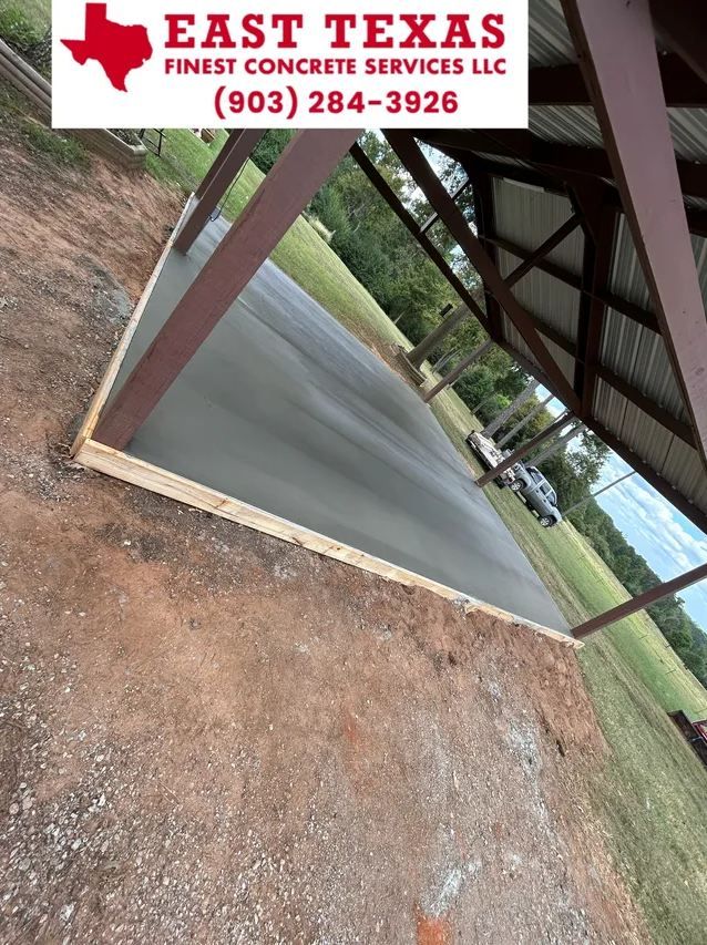 A newly poured concrete pad under a metal carport structure, framed by wooden forms, outdoors on a sunny day.