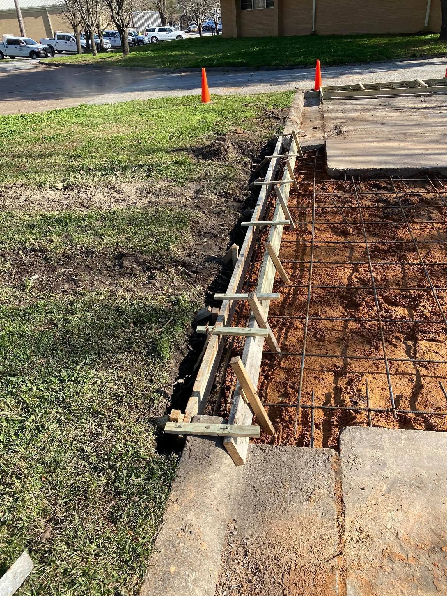 Wooden construction forms and rebar prepared for pouring concrete next to a grass verge and an existing pavement.