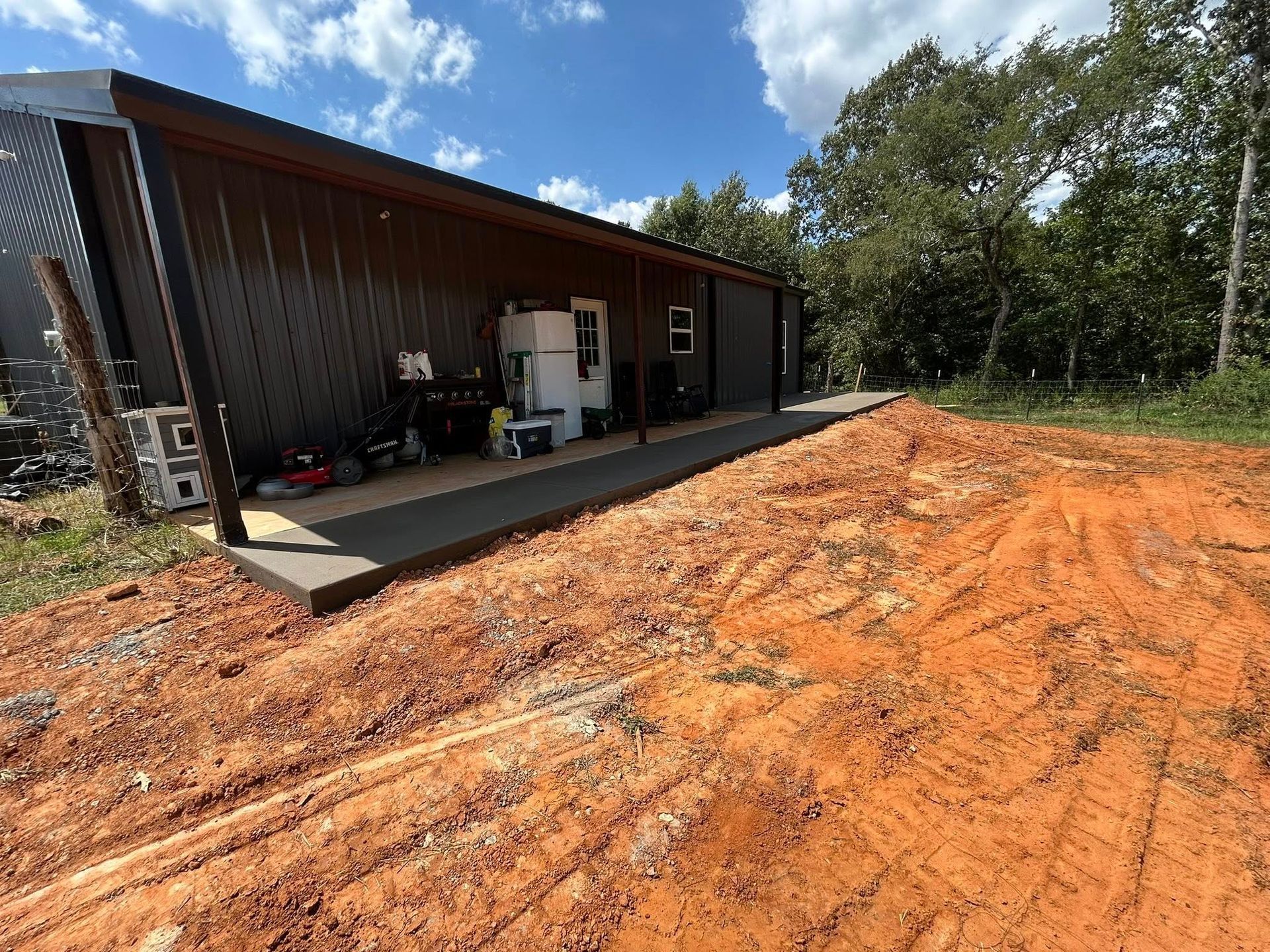 A dark metal building with a covered patio, featuring a refrigerator and outdoor gear, beside a patch of red dirt.