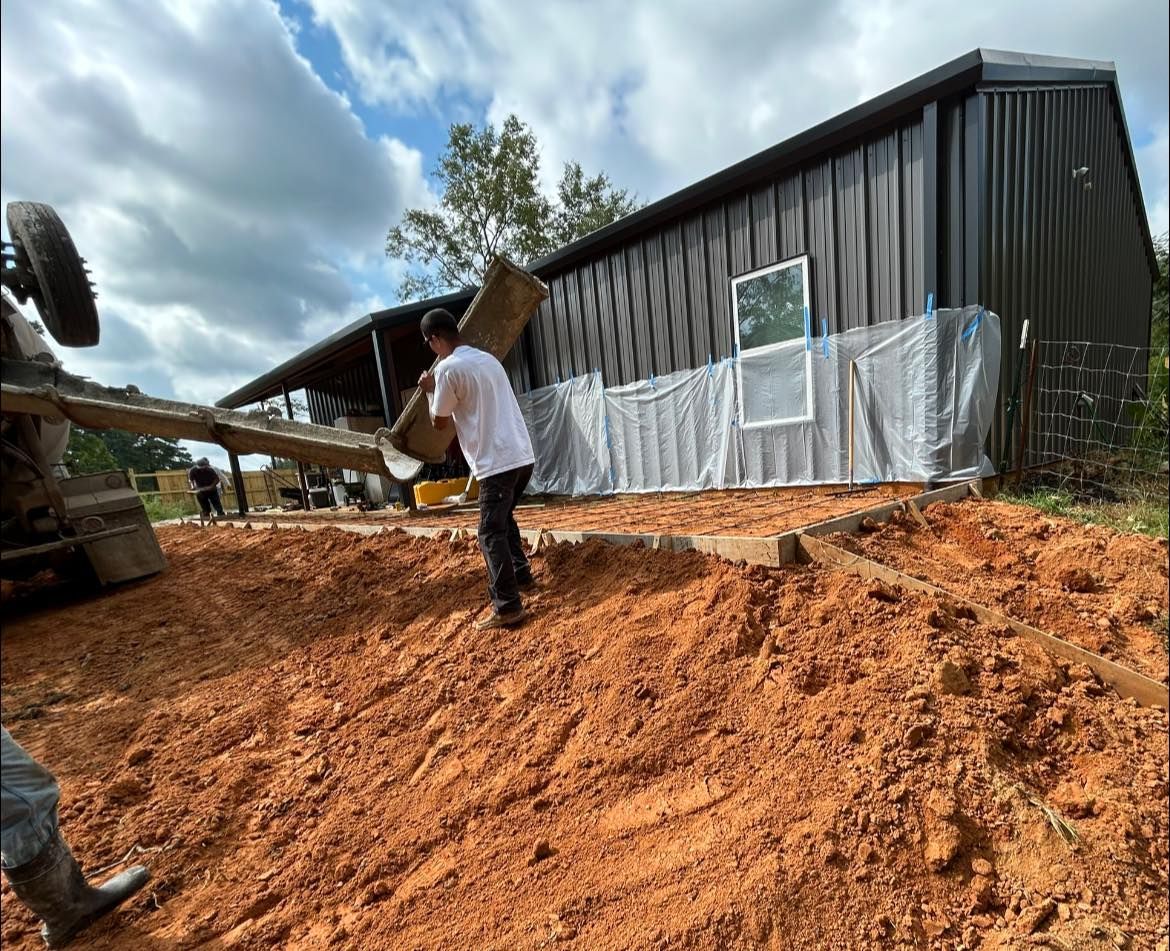 A worker guides a concrete chute from a truck toward a wooden frame beside a dark, metal-sided building on a red dirt site.