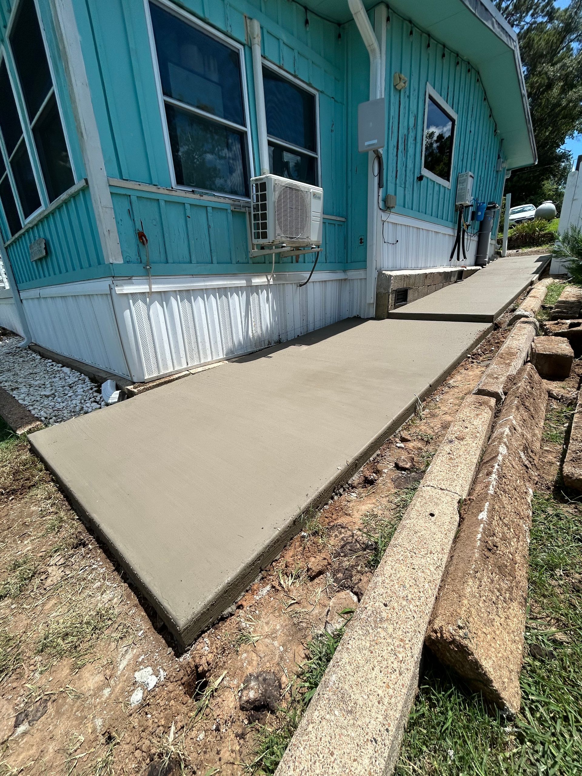 A new, smooth concrete walkway leads up to the side entrance of a turquoise mobile home.