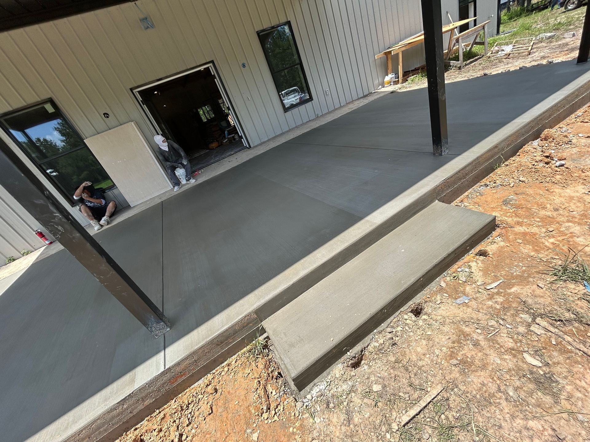 A fresh concrete patio with a single step under construction, next to a building with metal siding and a dark door.