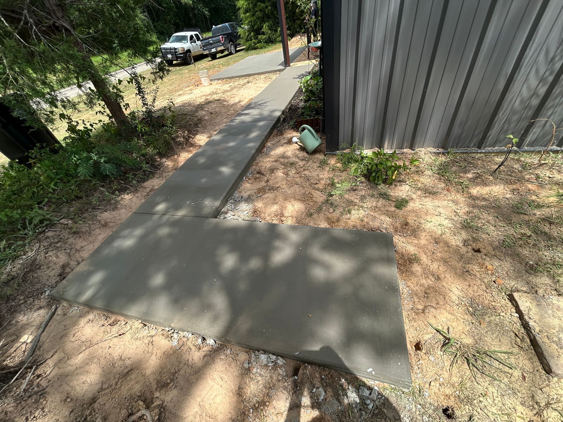 A new L-shaped concrete sidewalk leads from a driveway toward the side of a metal building on a dirt-covered yard.