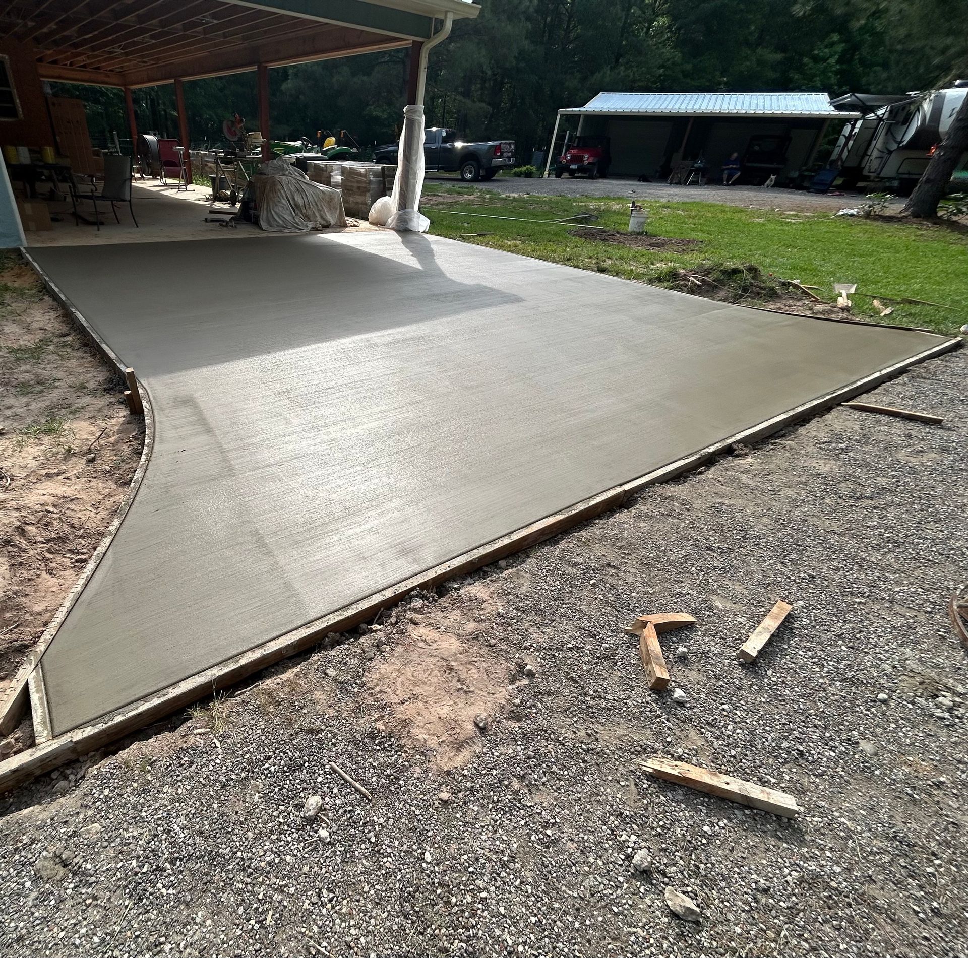 A freshly poured concrete patio surface extends from under a wooden porch roof, bordered by wooden forms and gravel.