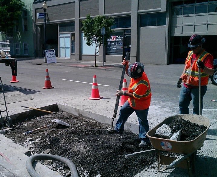 Two road workers in high-visibility vests and helmets work at a construction site on a paved city street.