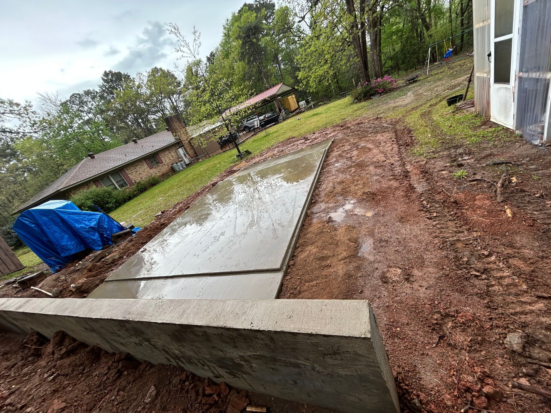 A freshly poured concrete patio slab under construction next to a low retaining wall in a muddy, sloping backyard.