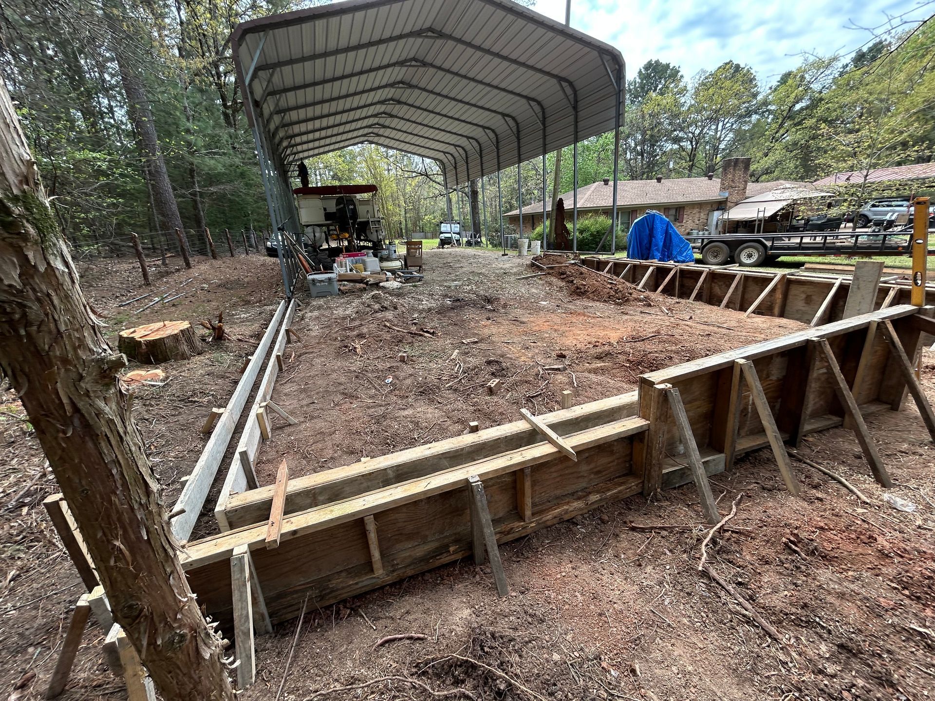 Wooden formwork for a concrete foundation being prepared in a wooded area under a metal carport structure.