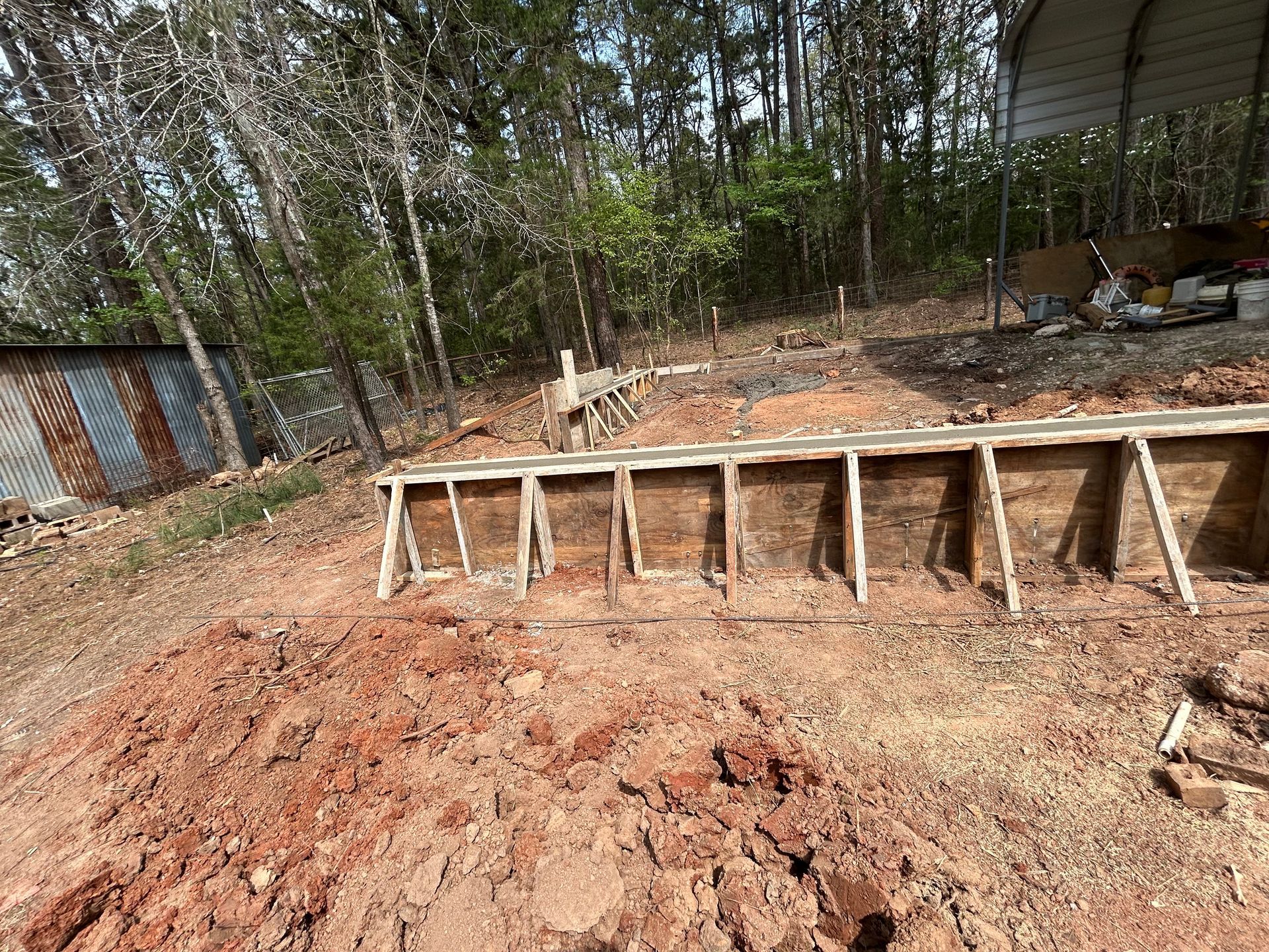 Wooden formwork for a concrete foundation wall set in a dirt lot with a forest background.