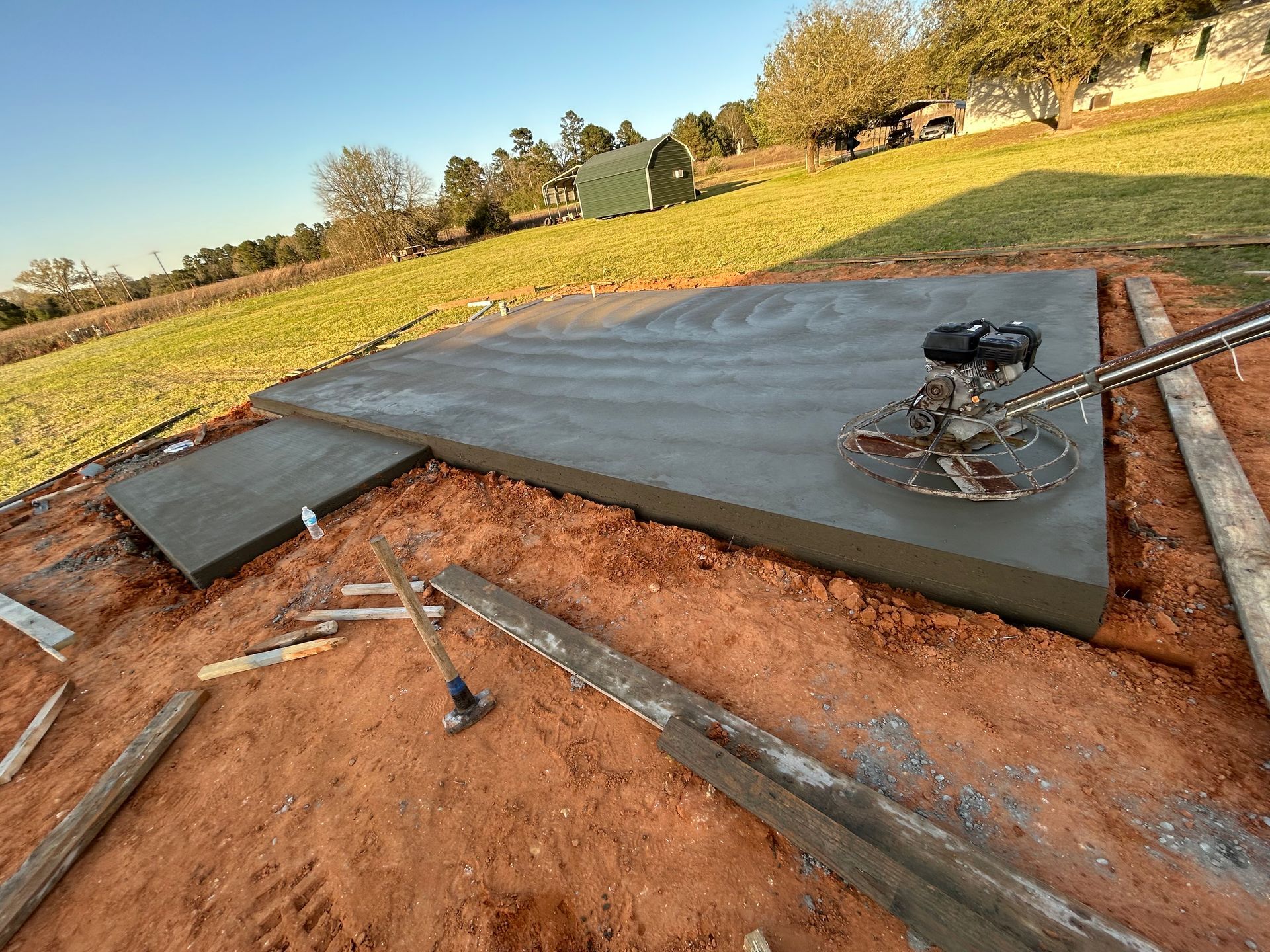A fresh concrete slab outdoors with a power trowel machine sitting on top, surrounded by red dirt and wooden forms.