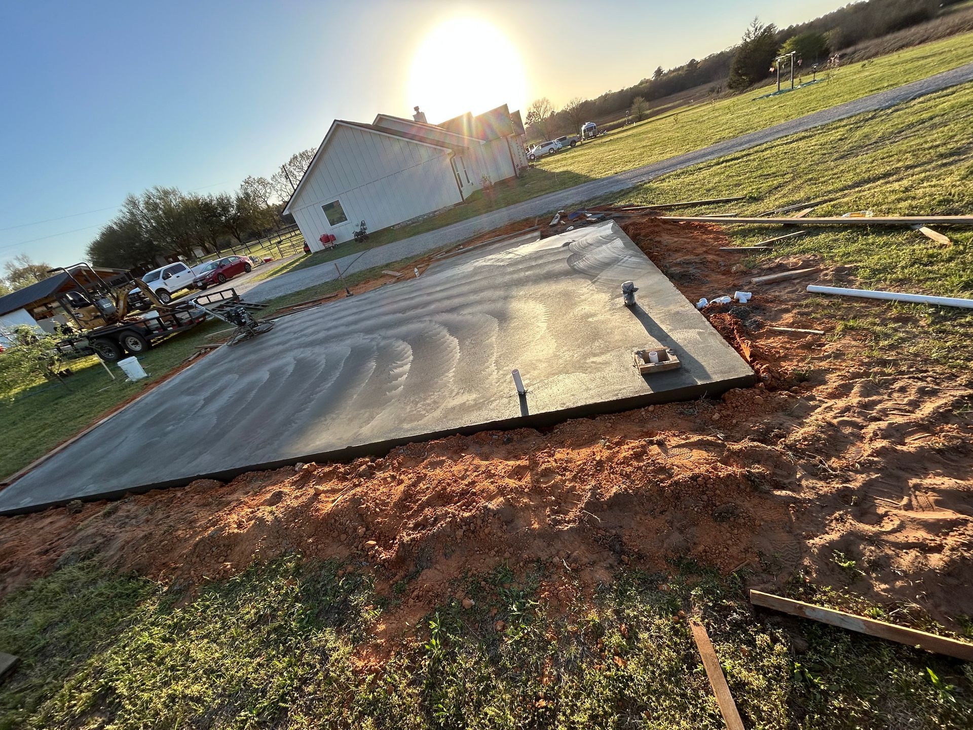A freshly poured concrete slab sits in a grassy yard under a bright, sunny sky near a house.