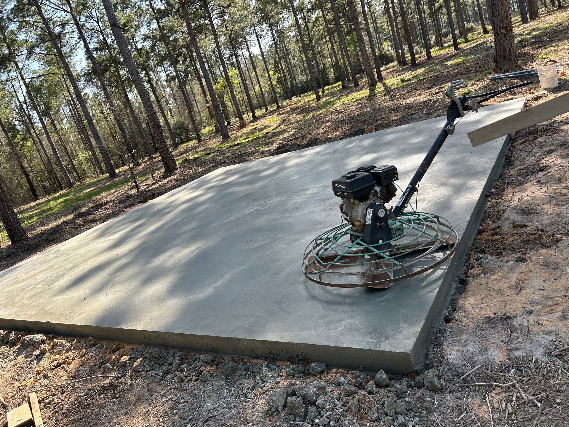 A power trowel sits on a freshly poured concrete slab in a wooded area.