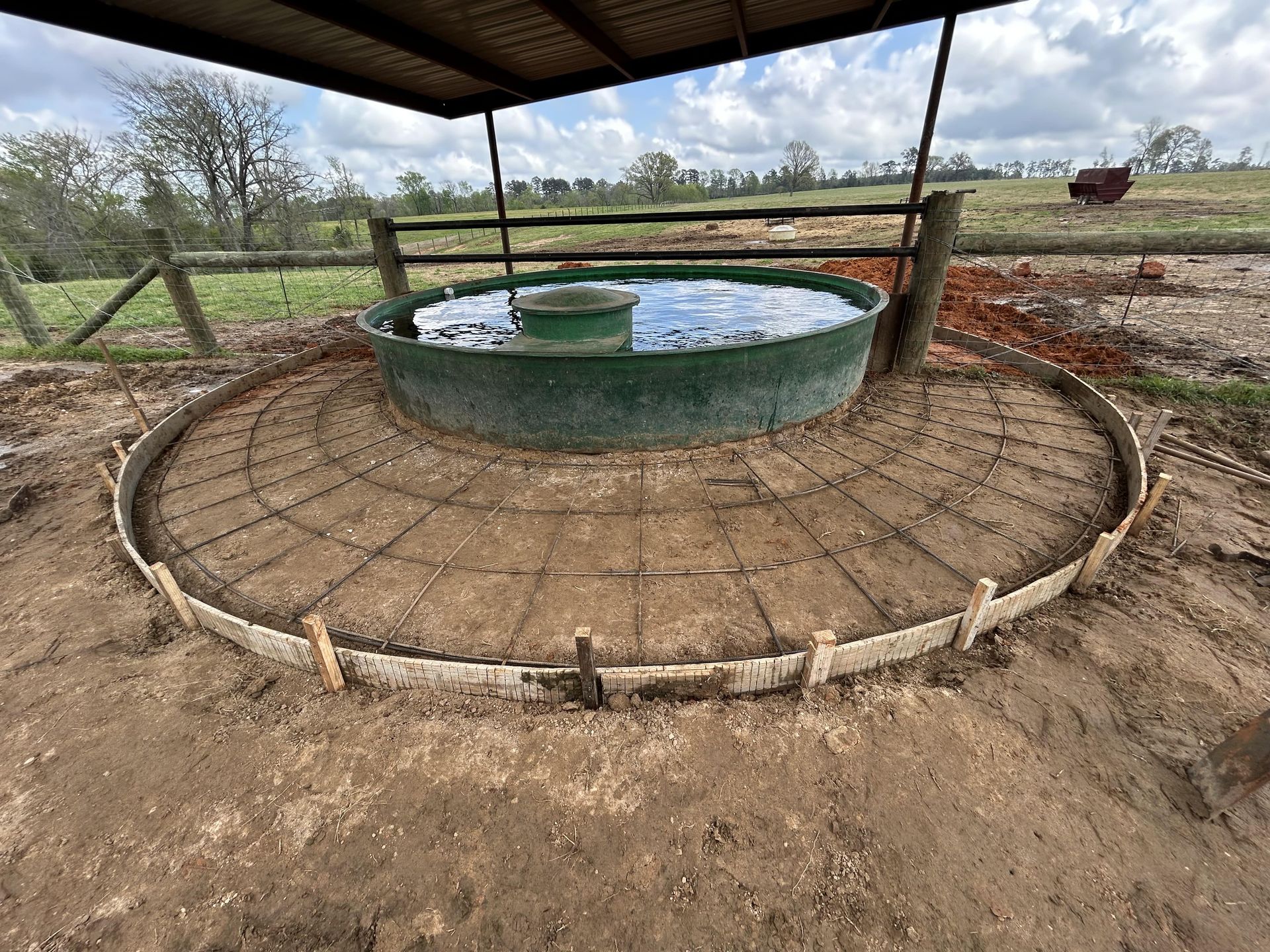 A green circular livestock water trough surrounded by metal wire reinforcement mesh inside a wooden concrete form.