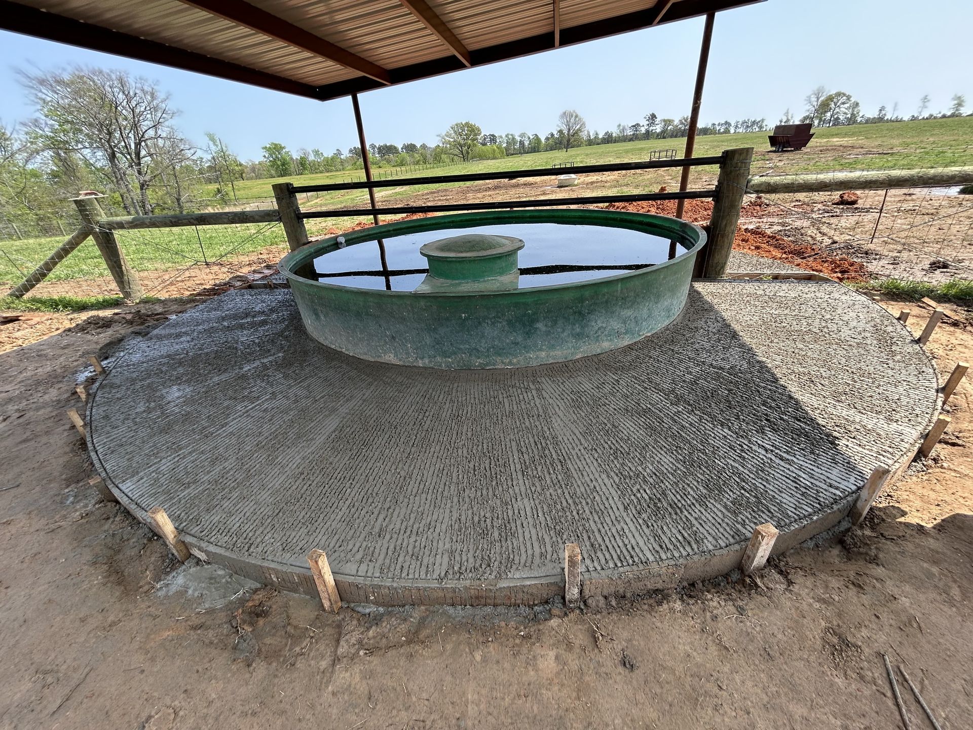 A circular water trough sits in the center of a freshly poured, semicircular concrete pad under a metal shelter.