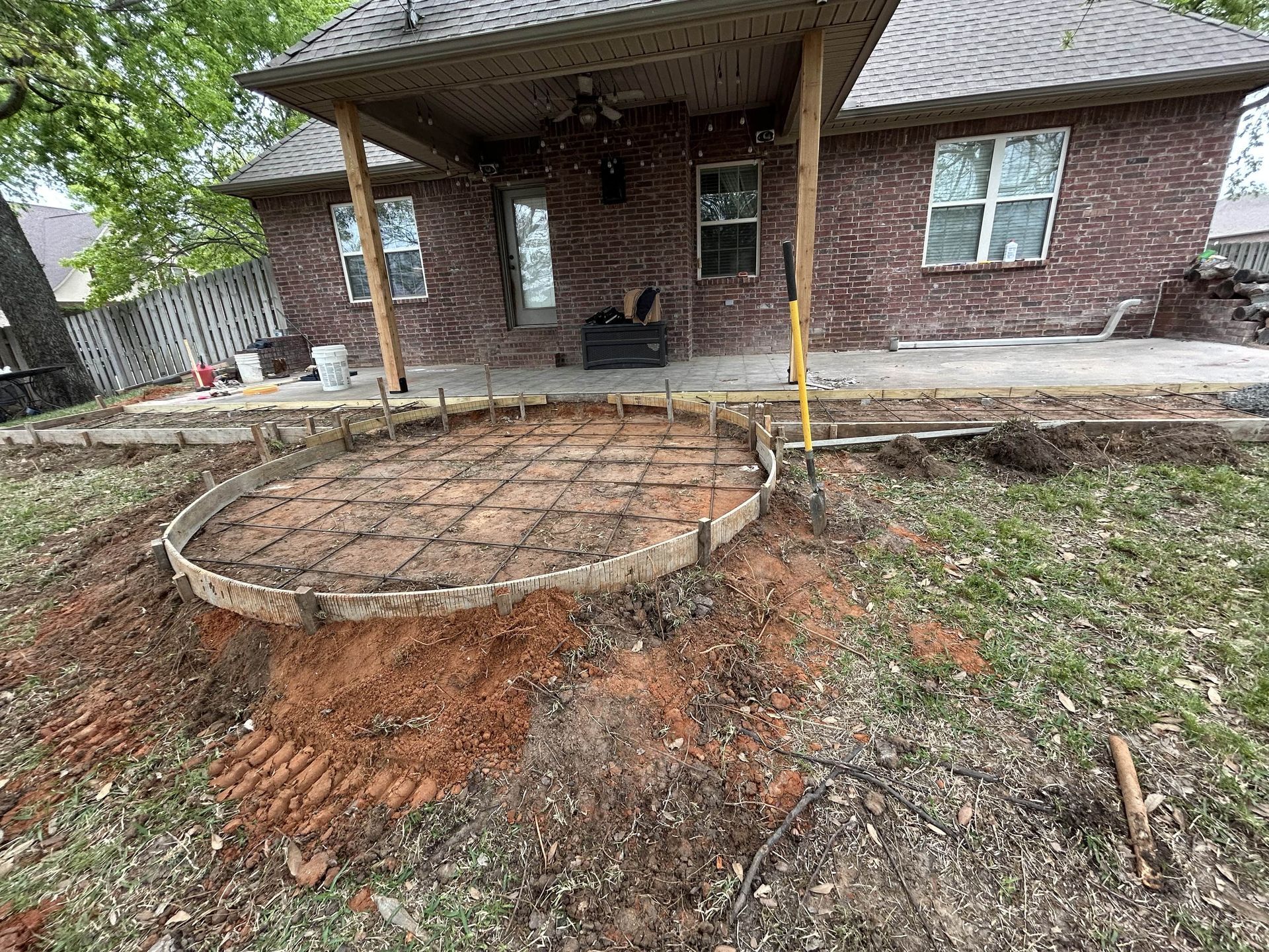 A circular concrete patio form with metal reinforcement mesh sits in a backyard in front of a brick house.