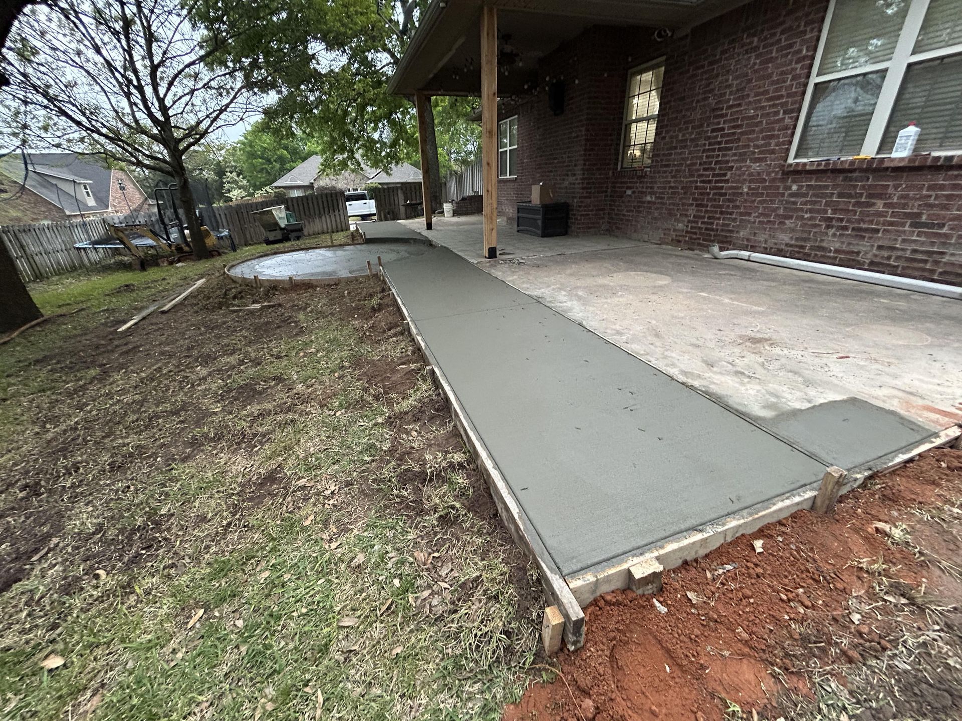 A newly poured concrete walkway extends from a brick house patio across a dirt and grass yard.