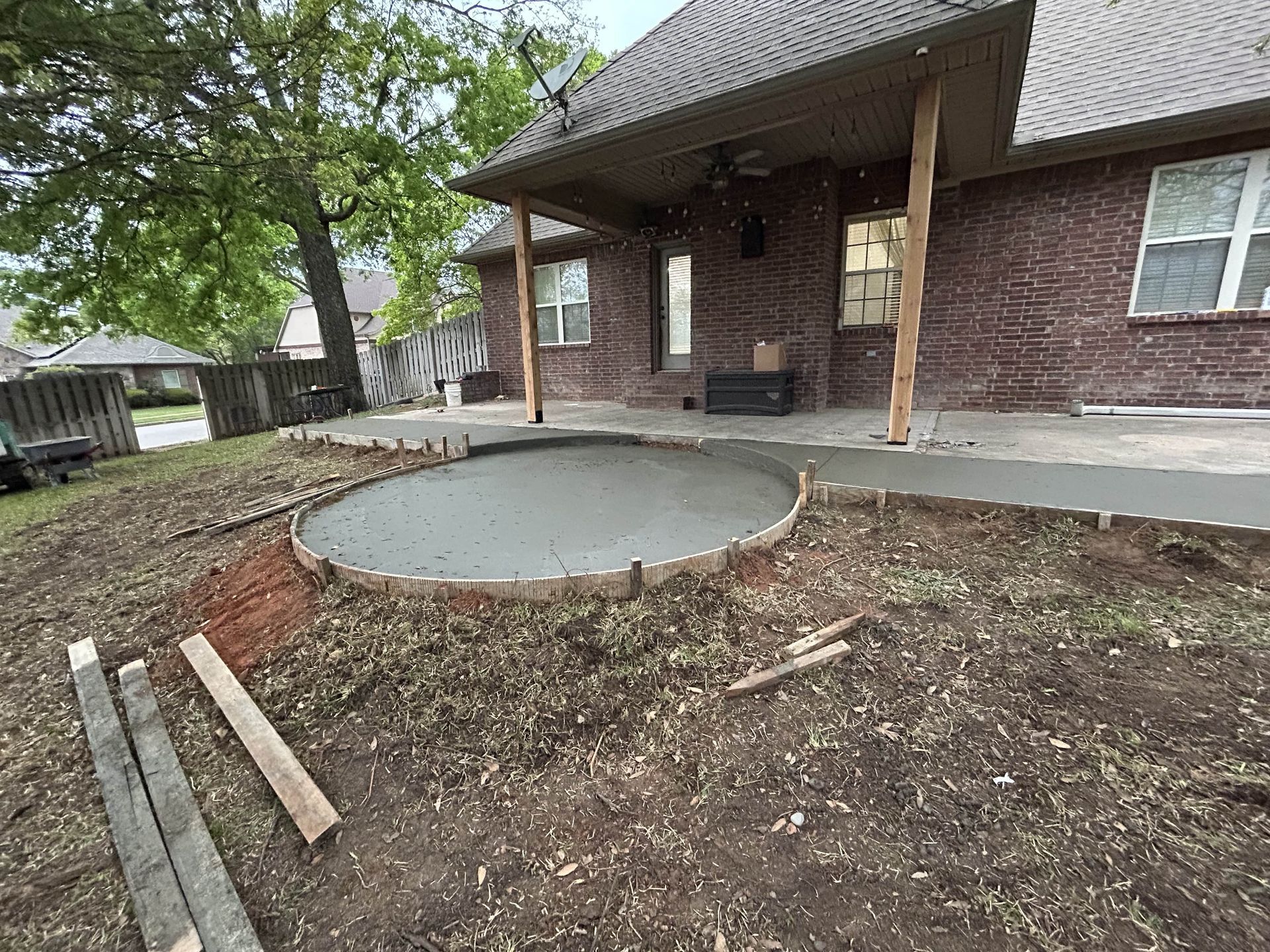 A newly poured circular concrete patio pad set in a grassy backyard next to a brick house with a covered porch.